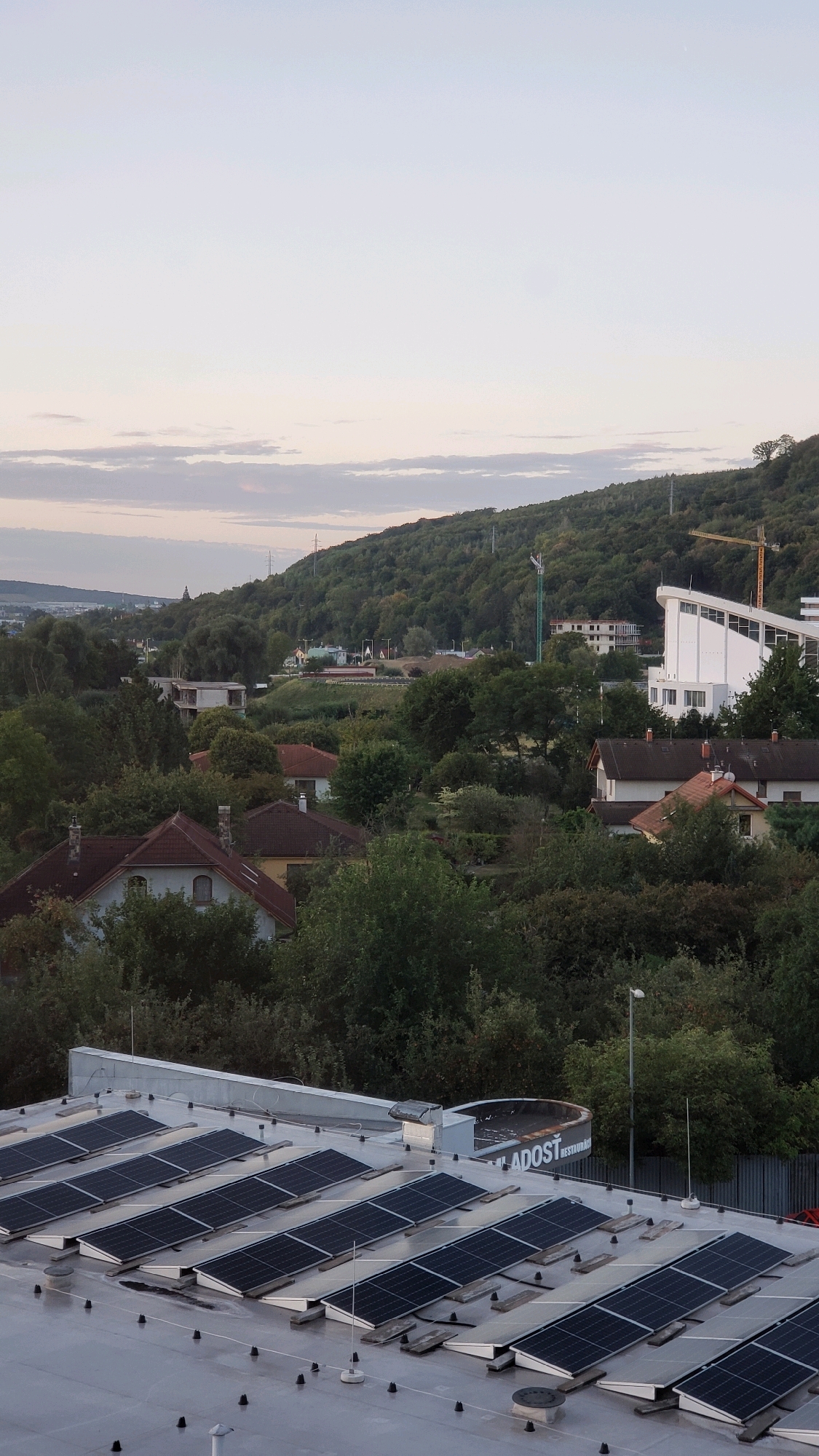 The image depicts a rooftop scene in Prešov, Slovakia, with a cityscape and a forested hill in the background. The foreground is dominated by a large, grey, flat roof covered in rows of solar panels. The panels are angled and arranged in neat, parallel lines, with some wooden supports visible beneath them. Several black, conical objects, likely roof anchors or mounts, are scattered across the roof surface. A white ventilation unit or air conditioner is mounted on the roof towards the top center. To the right of this unit, a partial view of a circular structure with signage is visible, including the partially obscured text "LADOST RESTAURAC".
Beyond the rooftop, the mid-ground reveals several residential houses with red-tiled roofs nestled amongst dense green trees. Further in the distance, a modern white building with a distinctive angular roofline stands out. The background is characterized by a lush, green, tree-covered hill that slopes upwards. The sky above is a pale blue with faint, wispy clouds, suggesting it might be early morning or late afternoon. Construction cranes, one yellow and one green, are visible on the hillside, indicating ongoing development. Power lines and poles are also present in the landscape. The overall impression is a suburban or edge-of-city view, blending residential areas, commercial structures, and natural topography.