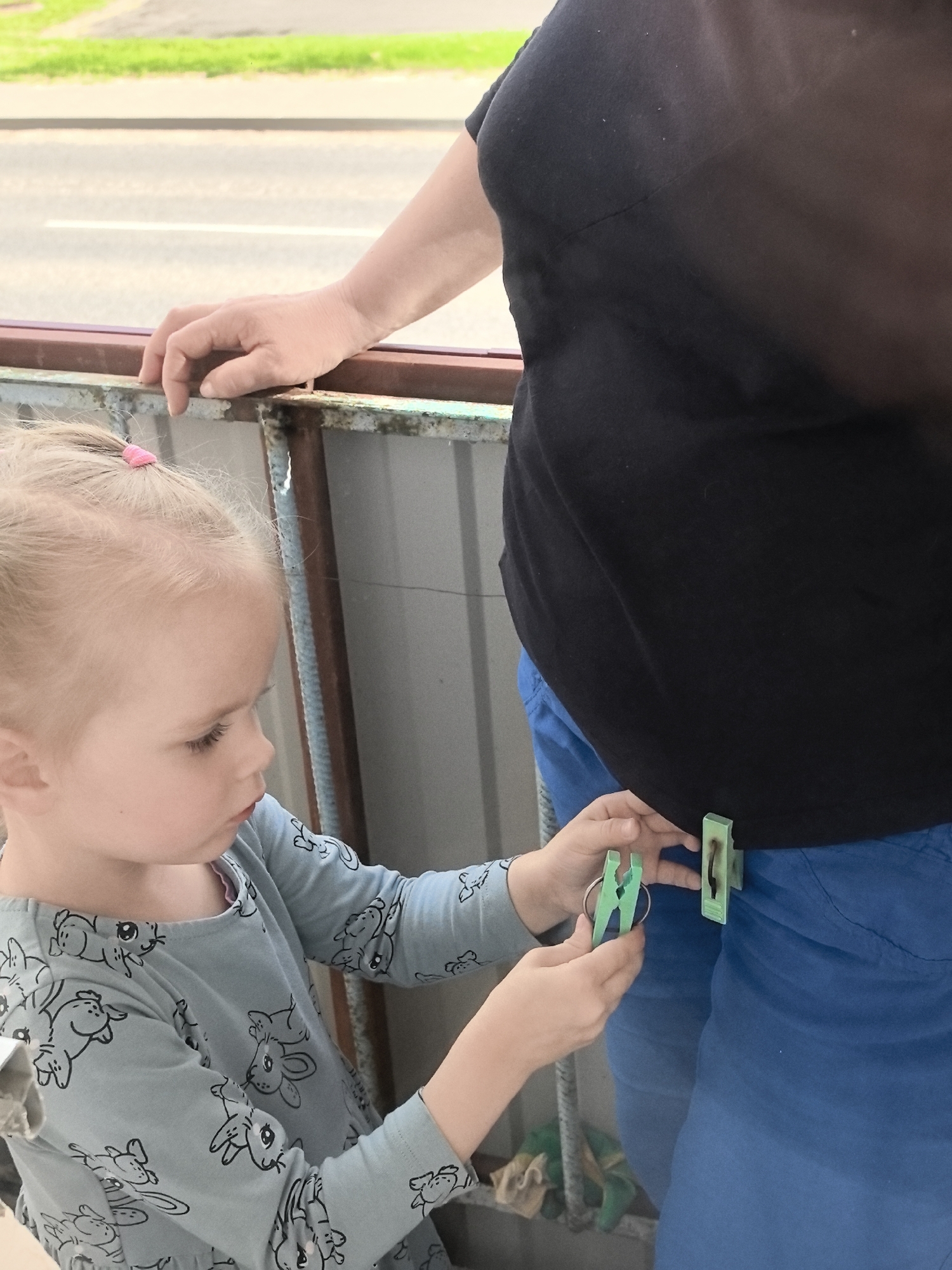 A blonde-haired young girl wearing a light blue long-sleeved shirt with a rabbit pattern is standing on a balcony and clipping two green clothespins onto the blue pants of a person wearing a black shirt. The girl is holding one clothespin in her left hand and is about to clip it onto the pants. The other person's right hand is resting on the balcony railing. The background shows a street with green grass and trees. The balcony appears to be made of metal with some rust and peeling paint.