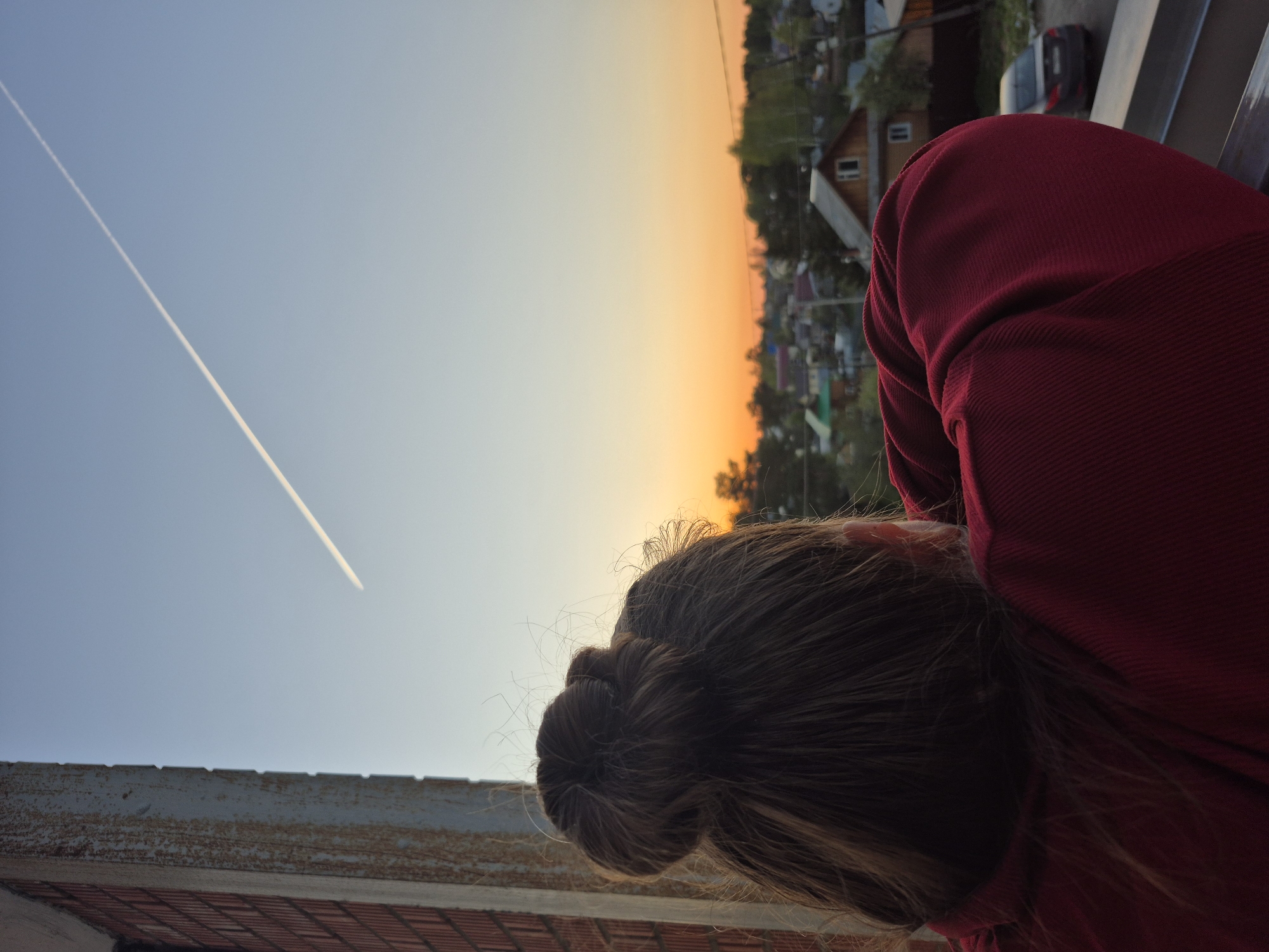A person with brown hair in a bun, wearing a red hoodie, leans over a balcony overlooking a town during sunset. The sky transitions from light blue to orange, with a contrail visible across the blue portion. In the background, small houses and greenery are visible, and a grey car is parked near a house. The balcony has a weathered, grey railing. It is likely this image was taken in the city of Chaikovsky, Russia, based on the context provided.