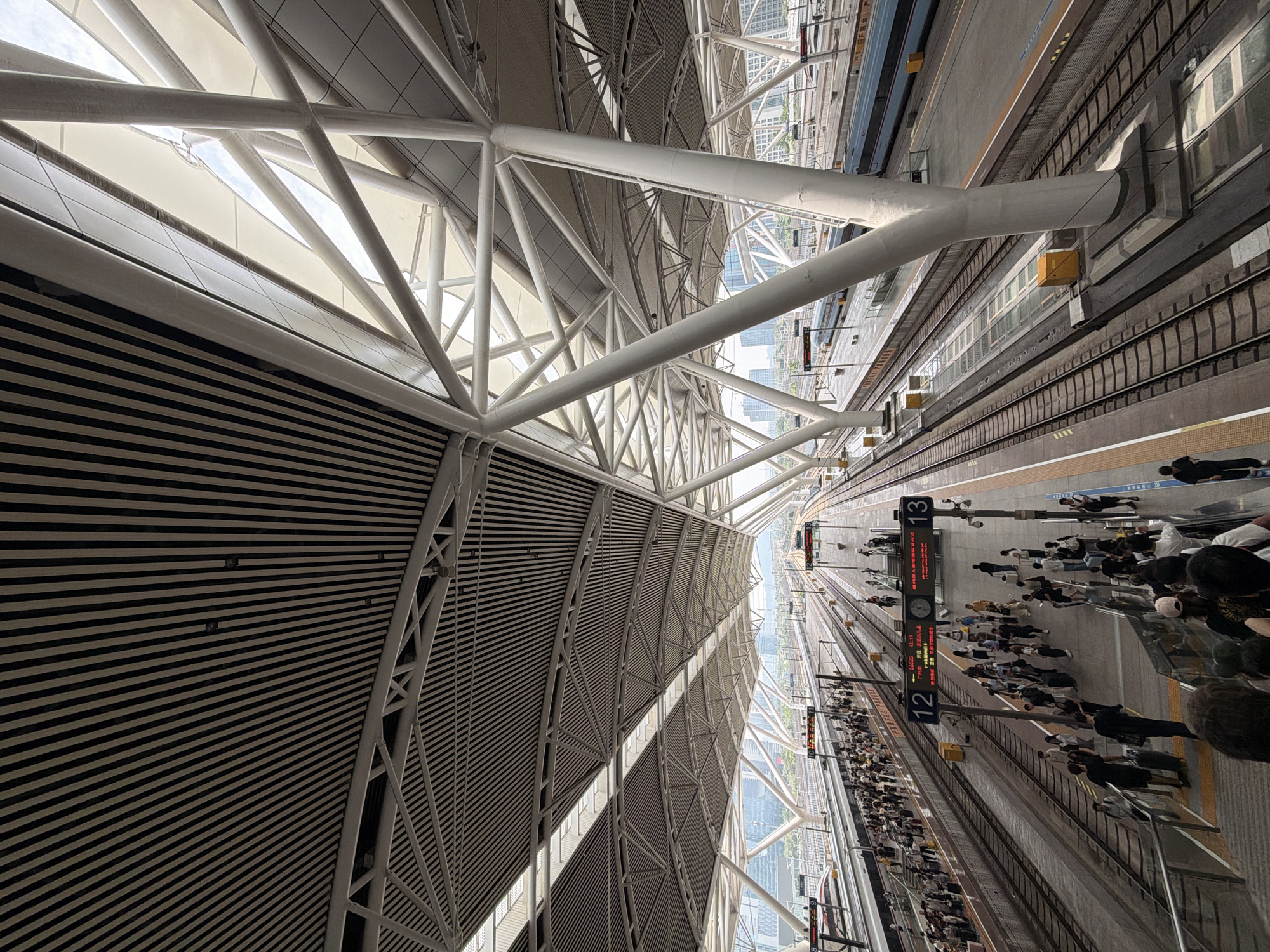 This is a photograph of the Guangzhou South Railway Station in Panyu District, China. The image captures the interior of the station's main concourse, focusing on the architectural design of the roof and the platforms below. The roof is a complex structure of white steel beams and supports, with sections covered in a textured, metallic material and others featuring translucent panels that allow natural light to filter through. The overall effect is modern and spacious, with a sense of an organized, albeit busy, environment.
Below the roof, multiple railway tracks stretch into the distance, flanked by elevated platforms. On these platforms, numerous people are visible, some standing in groups, others walking with luggage, and a few are interacting with each other. The people are dressed in casual clothing, indicating they are likely travelers. Digital information boards are positioned along the platforms, displaying departure and arrival times, further suggesting the active nature of a transportation hub. Trains are present on some of the tracks, their sleek designs hinting at high-speed rail. The environment appears to be clean and well-maintained, with a mix of natural and artificial lighting creating a bright atmosphere. The background shows glimpses of the urban cityscape with buildings reaching towards the sky, contextualizing the station within a developed metropolitan area.