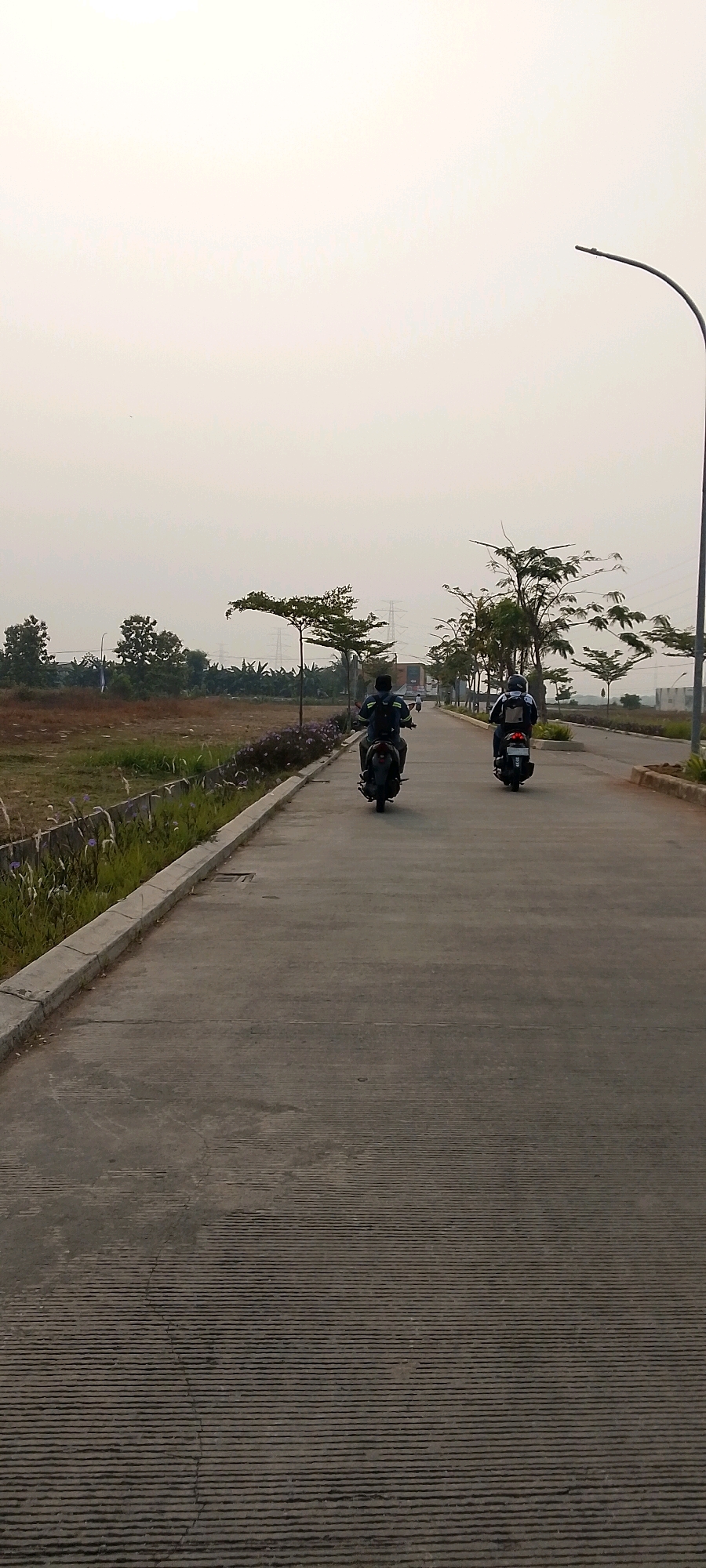 Two individuals are riding motorcycles on a paved road in Satriamekar, Indonesia. The person in the foreground is wearing a dark shirt and a black helmet, while the person behind them wears a dark shirt, a black helmet with white accents, and a backpack. Both motorcycles are dark in color. The road is a two-lane asphalt surface with visible horizontal grooving. On the left side of the road, there is a concrete curb and a strip of dry grass and small purple flowers. Beyond the vegetation, there is an undeveloped plot of land with brown soil and scattered vegetation. In the distance, several trees are visible, including some with a distinctive umbrella shape. Power lines and electrical towers are also present in the background, along with some buildings and structures. A street lamp is positioned on the right side of the road in the upper portion of the image. The sky is overcast and light in color, suggesting it may be early morning or late afternoon.
