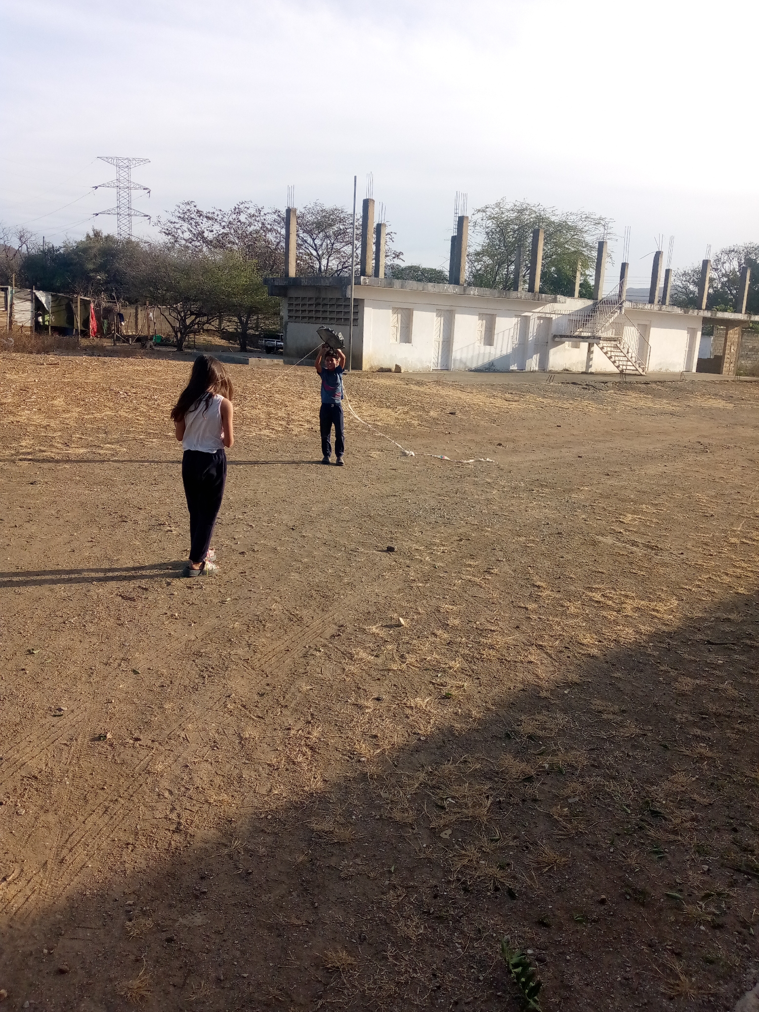 This media file depicts an open, dusty outdoor scene, likely in a semi-rural or undeveloped area of Juan Griego, Venezuela, under a clear sky. The ground is dry, light brown earth with sparse, dried grass and visible tire tracks, characteristic of arid or semi-arid conditions.
Two individuals, appearing to be children or young adolescents, are the main subjects. In the mid-ground, with her back to the camera, is a girl with long, dark hair, wearing a white tank top and dark pants. Her elongated shadow stretches towards the bottom right of the frame, suggesting the sun is low in the sky, indicating either early morning or late afternoon. Further back and slightly to the right, a boy faces the girl, holding a dark, possibly plastic, basin or tray above his head with both hands. He is dressed in a blue t-shirt and dark pants. A light-colored string or rope can be seen extending from his area towards the girl, implying they are engaged in an activity or game together.
The background features a large, white, partially constructed building with numerous exposed concrete pillars and rebar protruding from its roofline. A metal staircase is visible on the side of this structure. To the left of this building, a cluster of trees and shrubs partially obscure some smaller, makeshift structures or shacks. In the far distance, beyond the trees, a prominent metal electricity transmission tower stands against the light sky. The overall scene suggests a blend of informal settlement or developing infrastructure within a natural, dry landscape. There is no visible text in the image.