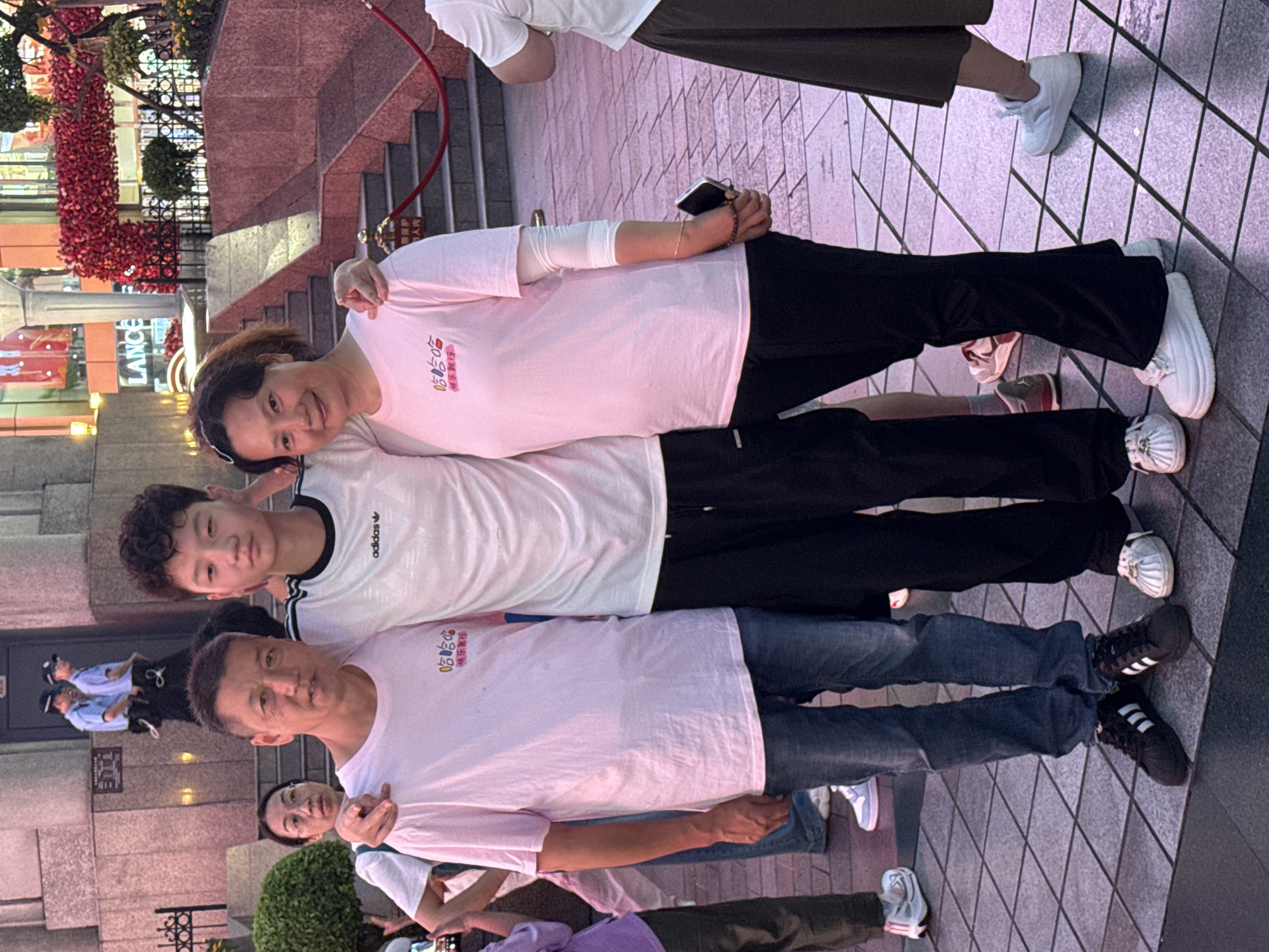 A family of three, a father, mother, and teenage son, pose for a photo outdoors at street level. The father, on the left, is wearing a pink t-shirt and jeans, with his arm around his son. The mother, on the right, is also wearing a pink t-shirt and black pants, with her arm around her son. The son is in the center, wearing a white t-shirt and black pants. In the background, a staircase leads up to a building with red floral decorations and a sign for "LANCE." Two police officers are visible in the distance near the entrance of the building. The ground is paved with square tiles. The lighting suggests it is evening.