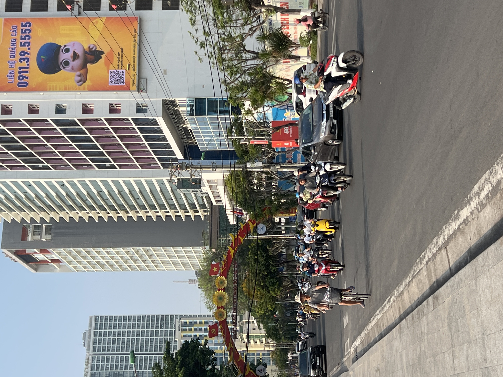 The image captures a bustling street scene in Nha Trang, Vietnam. Numerous motorbikes and scooters dominate the roadway, interspersed with a few cars. The sidewalks appear to be made of concrete. People are riding the motorbikes, some wearing helmets. Overhead, an ornate archway decorated with red flags and sunflower motifs spans the street. Tall buildings line the background, including one with a large advertisement featuring a cartoon character and Vietnamese text; the sign includes the phone number 0911.39.5555. The sky is clear. Utility poles and wires run along the street. The image is rotated clockwise.