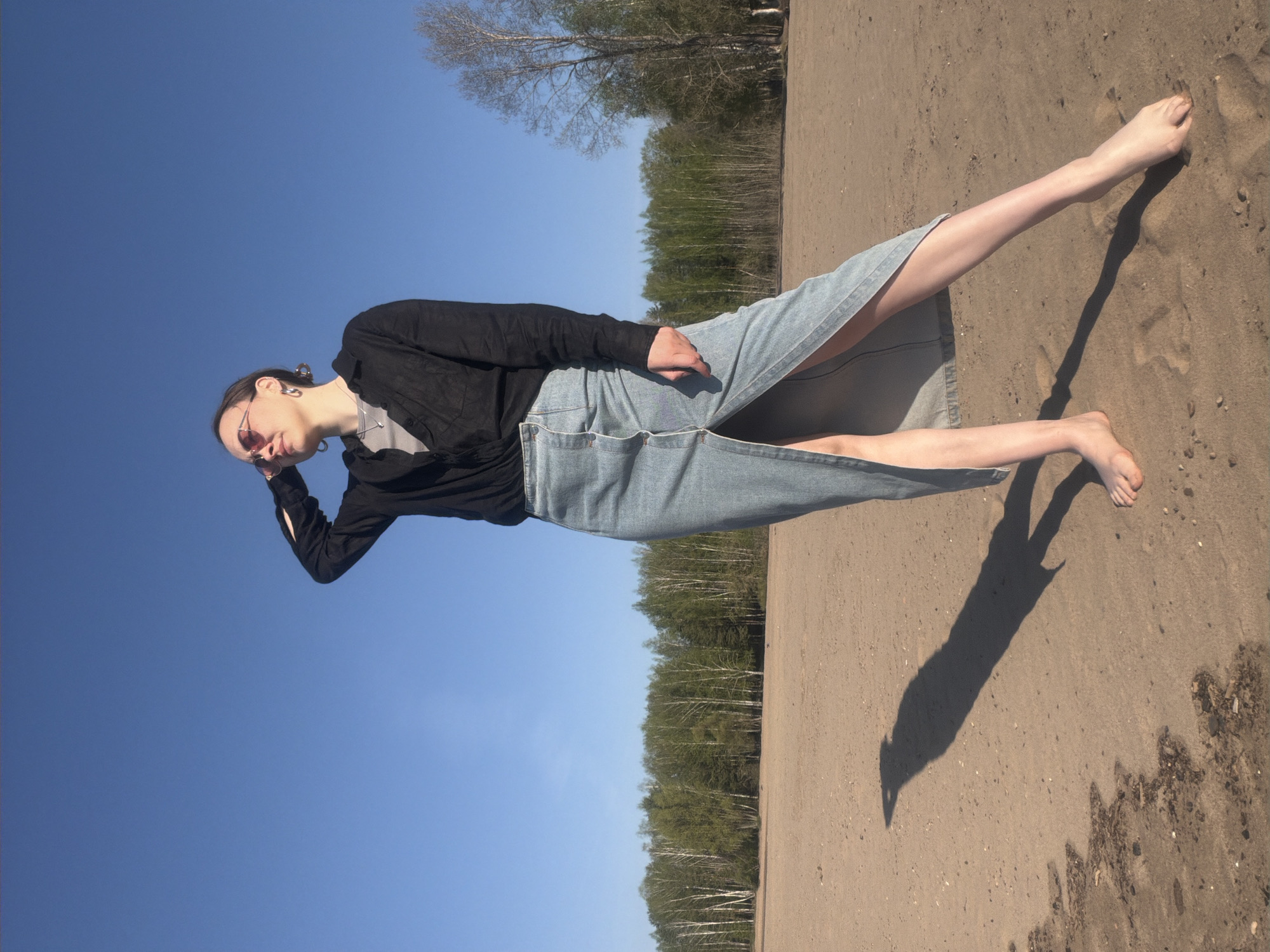 The photograph depicts a woman reclining on a sandy surface under a clear blue sky. She wears a black long-sleeved top, a grey t-shirt, and a light blue denim skirt with a slit. Her right leg is extended towards the top-right of the frame, and her left leg is bent. She has sunglasses on and is touching her forehead with her left hand. The sand appears slightly damp near the bottom-right corner of the image. In the background, a dense line of trees is visible along the horizon. Based on the location provided, Noviy, Russia, this appears to be a beach or sandy area near the shoreline.