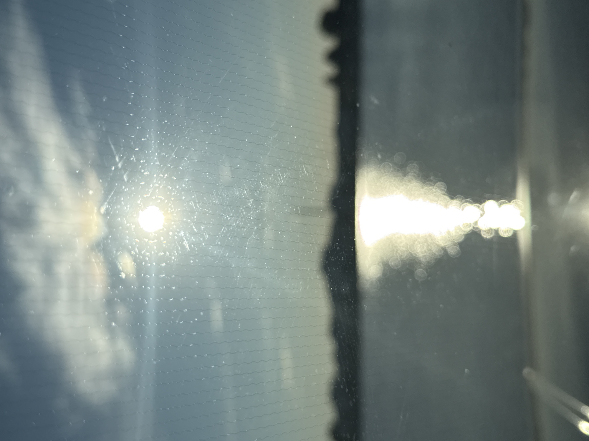 A bright sun shines through a hazy sky, casting a shimmering reflection on the water below. The sun's rays are diffused by what appears to be a gridded surface, possibly a window or screen, with small water droplets or smudges scattered across it. The horizon is blurred, showing a dark silhouette of land or buildings against the bright sky. The water itself is calm, with the reflection of the sun creating a luminous pathway across its surface. The overall impression is a serene, atmospheric view, likely captured from a vantage point looking out at a body of water.
