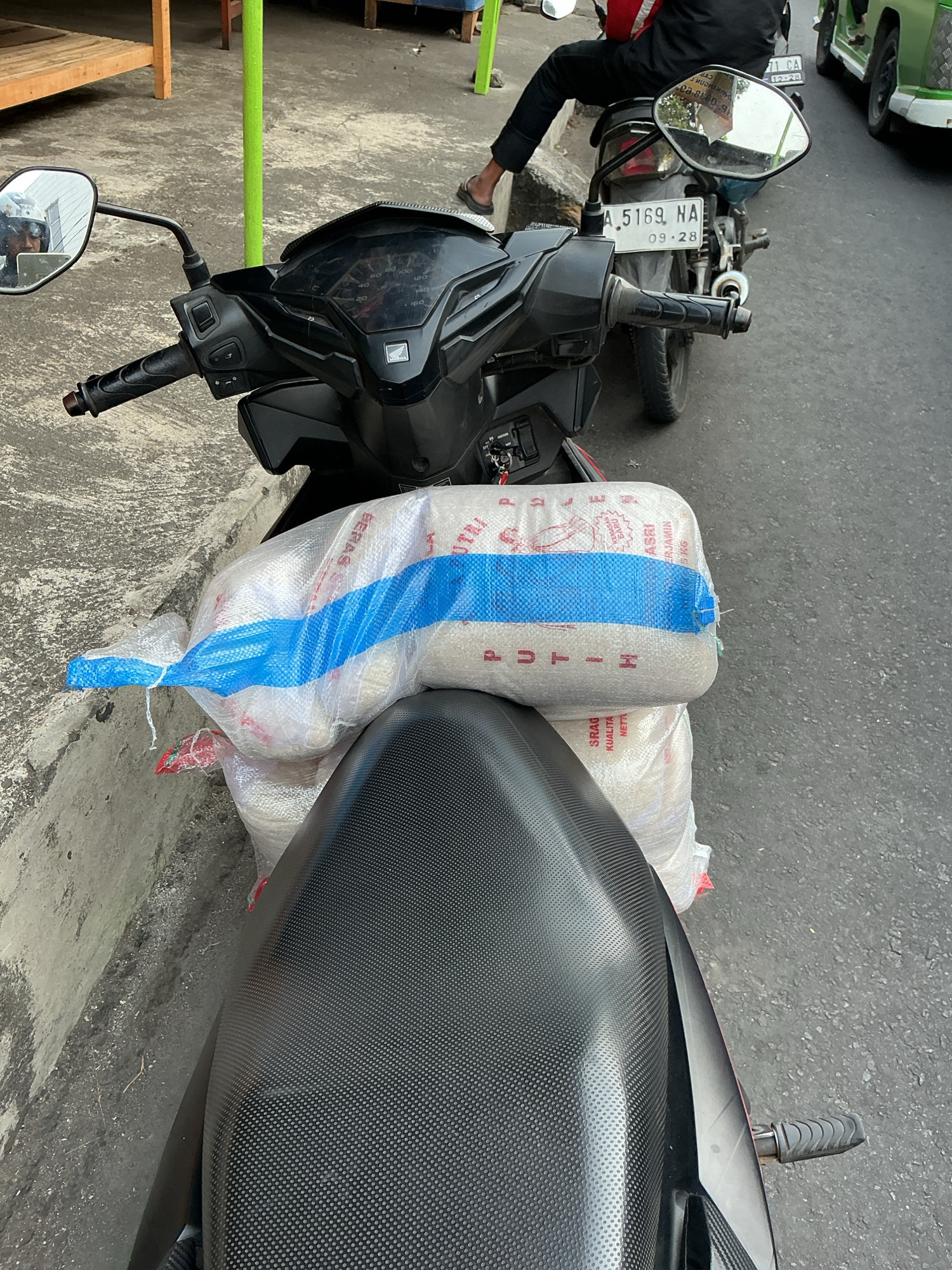 A POV shot from a motorcycle in Magelang, Indonesia shows two large sacks of rice tied to the rear. The motorcycle's handlebars and dashboard are visible in the foreground, with a reflective mirror showing the rider's helmet and a glimpse of the surrounding area. In the background, a person is seated on another motorcycle, facing away from the camera, with a green utility vehicle to their right. The ground is a mix of concrete and asphalt.
