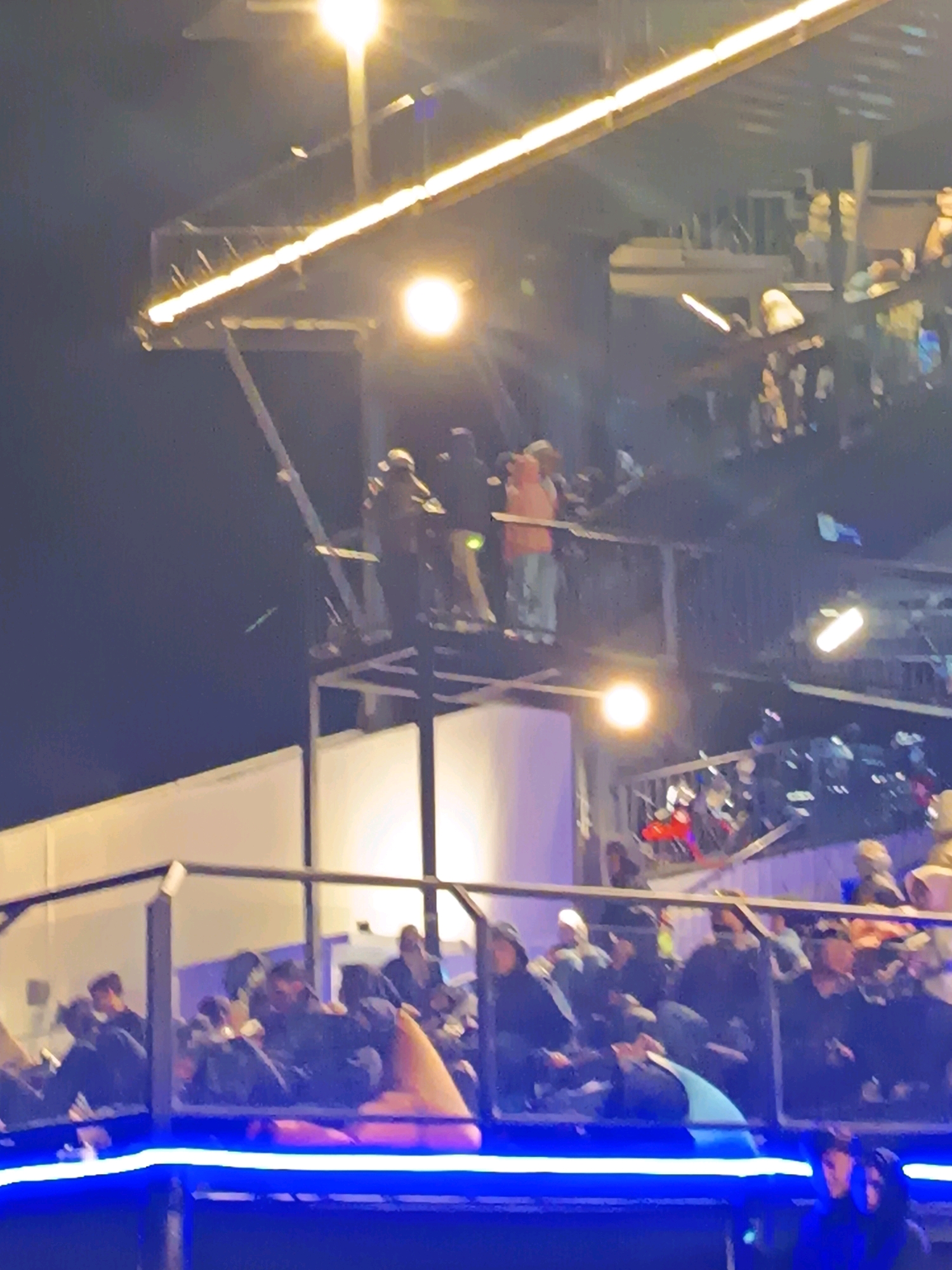 The image depicts an outdoor event at night, possibly a concert or festival, with multiple levels of viewing areas filled with spectators. Bright lights illuminate the scene, including long, horizontal light strips and circular spotlights. In the upper portion of the image, a group of individuals stands on a raised platform, some appearing to be interacting with each other. Below them, another tiered seating area is visible, densely populated with people seated and standing. The overall atmosphere is one of a lively gathering. The location is specified as Keditan, Indonesia.