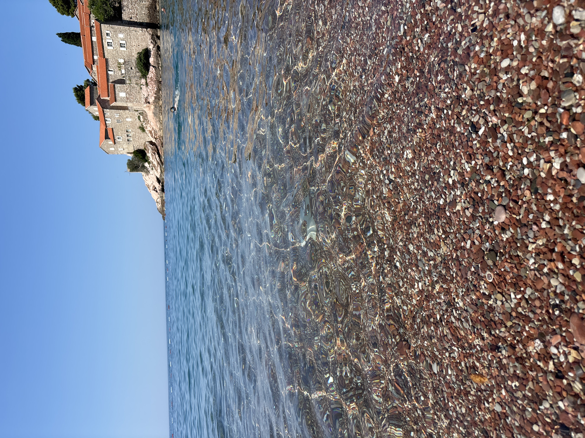 The image depicts a view of Sveti Stefan, a small islet and fortified village in Montenegro, from a pebble beach. The foreground is dominated by a dense layer of small, multi-colored pebbles, mostly reddish-brown with white and gray interspersed. These pebbles are partially submerged in clear, shallow water, with the sunlight creating intricate patterns of light and shadow on them.
Beyond the pebble beach, the water transitions to a deeper blue. The surface of the sea is gently rippled, and a line of white buoys stretches horizontally across the middle ground, marking a swimming area.
In the background, the historic island settlement of Sveti Stefan rises from the rocky coastline. It is characterized by stone buildings with characteristic red-tiled roofs, nestled on a rugged, tree-covered hill. A few cypress trees stand tall on the island.
A lone swimmer is visible in the water between the buoys and the island, their head and shoulders breaking the surface as they swim towards the island. The sky above is a clear, vibrant blue, indicating a sunny day. The overall scene conveys a sense of tranquility and natural beauty with a touch of historical charm.