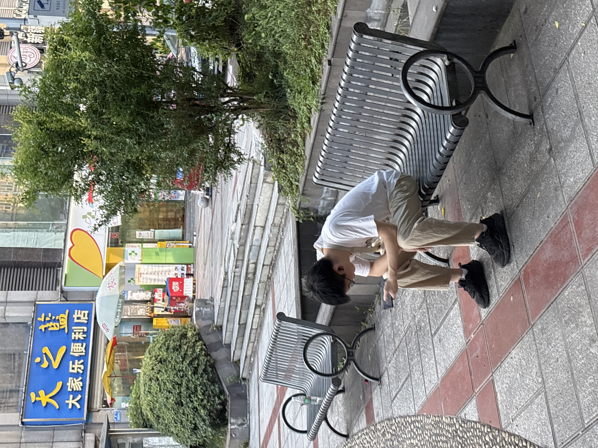 A young man wearing a white t-shirt, khaki pants, and black sneakers sits on a slatted metal bench outdoors, looking down at his black smartphone. He is wearing earbuds and appears to be focused on his device. To his left, another empty metal bench is visible, with a clear plastic bottle of water on it. The benches are situated on a paved walkway with a patterned tile design of red and gray squares. In the background, there are stone steps leading up to a building, and a lush green bush is positioned on the left side of the steps. A small, leafy tree grows to the right of the steps, with more dense foliage behind it. A blue and yellow sign with Chinese characters is mounted on the building above the bush. The overall scene suggests a public urban space, possibly in a city like Liangjiang, China, given the signage.