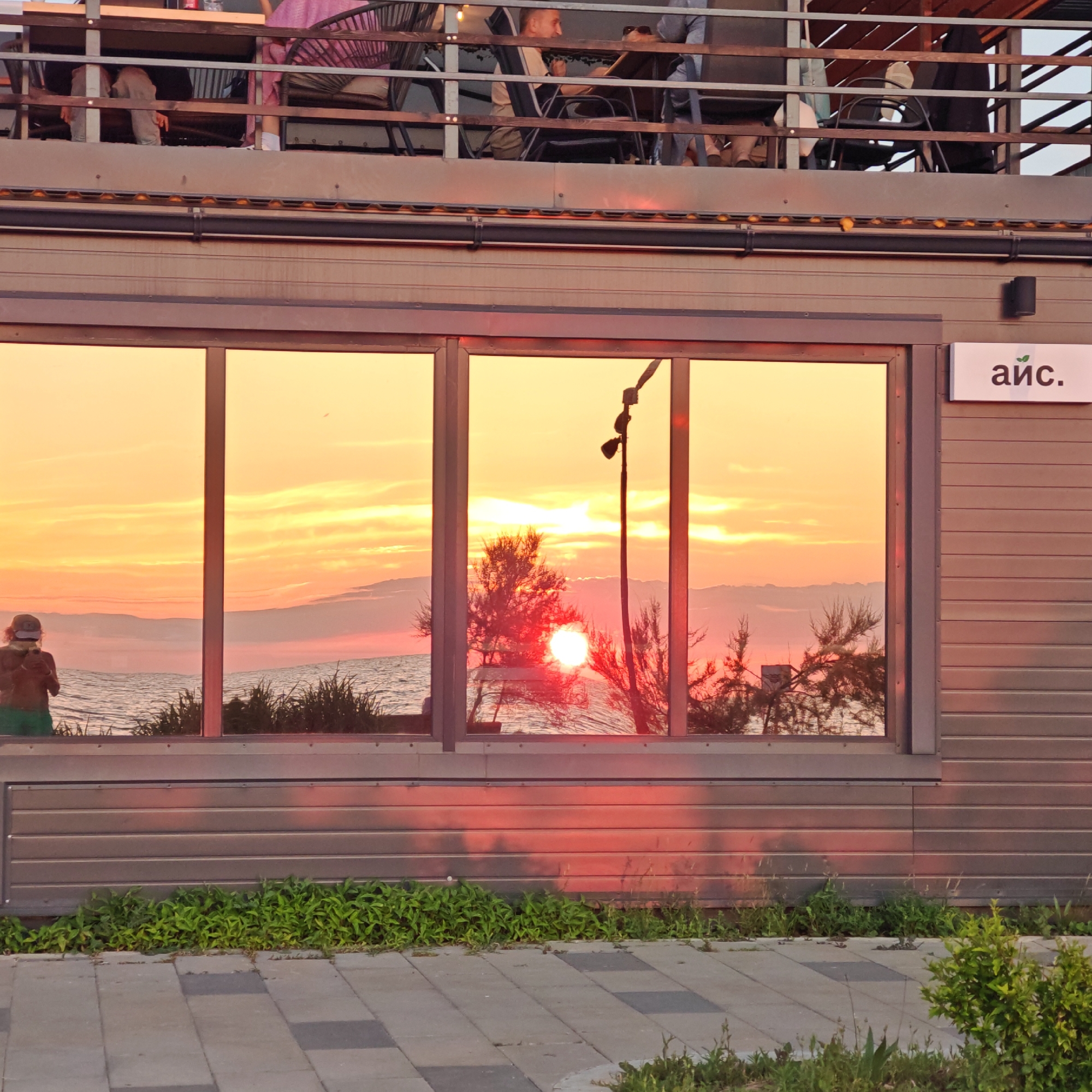The image shows a reflective window of a building. The window reflects a sunset over the ocean. In the foreground, there is a sidewalk and some greenery. The building itself is made of horizontal siding. A sign with Cyrillic letters is visible on the side of the building. The overall scene is calm and serene, with the warm colors of the sunset dominating the reflection.