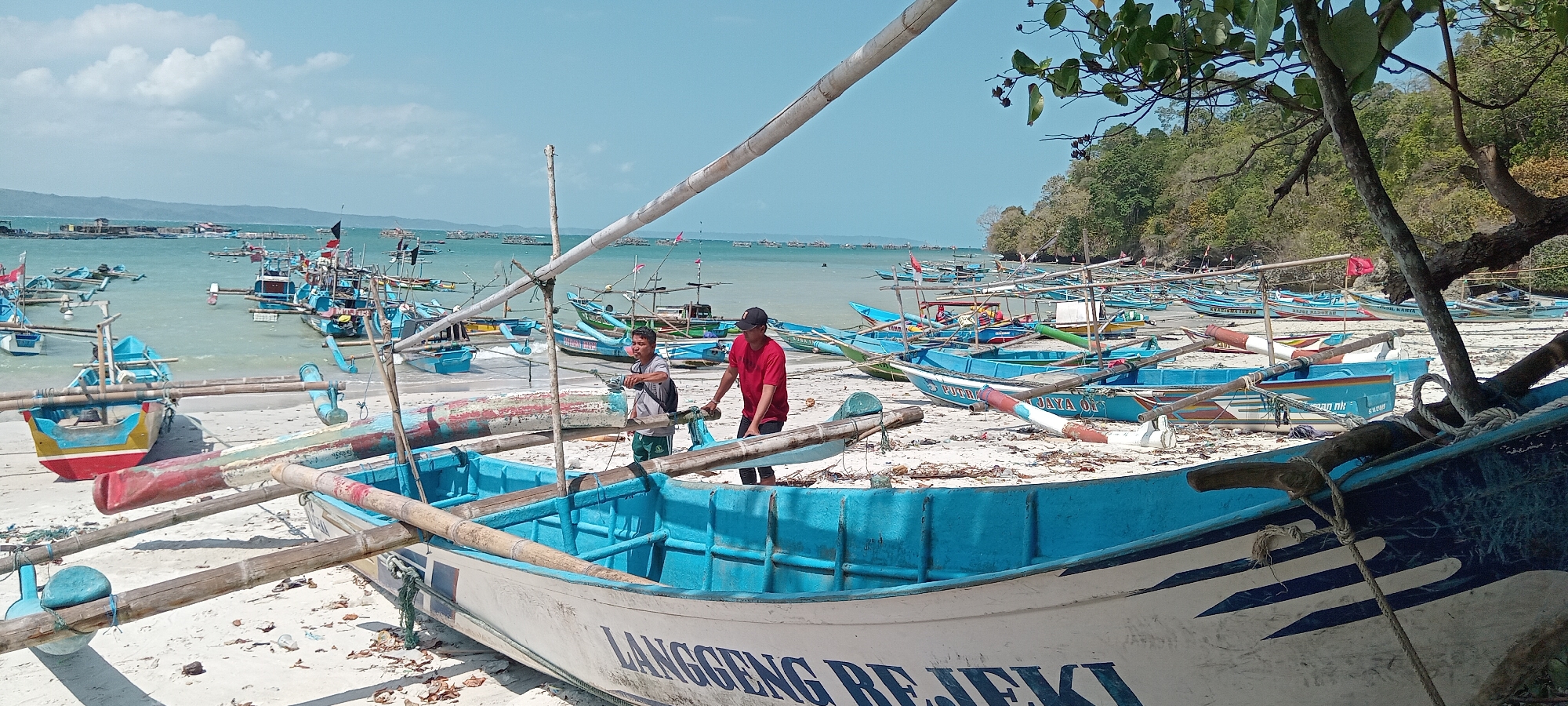The image depicts a crowded beach scene in Desa Pagandaran, Indonesia, filled with numerous traditional fishing boats. In the foreground, a white and blue boat with the inscription "LANGGENG REJEKI" is prominent, resting on the sandy shore. Two men are visible near this boat; one in a grey shirt and green shorts is looking towards the water, while another in a red t-shirt and black cap is standing with his hands on the boat. Behind them, a multitude of blue and other colored fishing boats are lined up on the beach and in the shallow, turquoise water. Some of these boats have flags and elaborate outriggers. In the background, a lush green hillside with trees overlooks the bay, and more boats are scattered in the distance, suggesting an active fishing community. The sky is bright blue with scattered white clouds. The beach appears to be composed of light-colored sand with some debris and dried leaves.