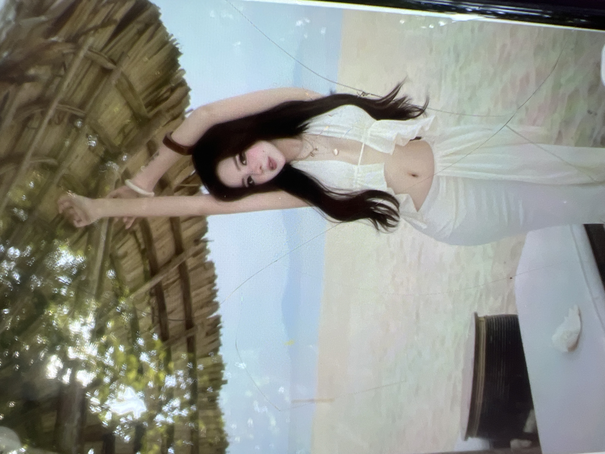A young woman with long dark hair and fair skin, wearing a white halter top and a white sarong, poses with her arms raised under a thatched umbrella on a sandy beach. The background shows a light blue sky and a hazy body of water. In the foreground, a white lounge chair with a dark cylindrical object beside it is visible. The image is a photograph, and the scene is likely a beach resort. The location in Kara-Dıykan, Kyrgyzstan is not supported by any visual evidence in the image.
