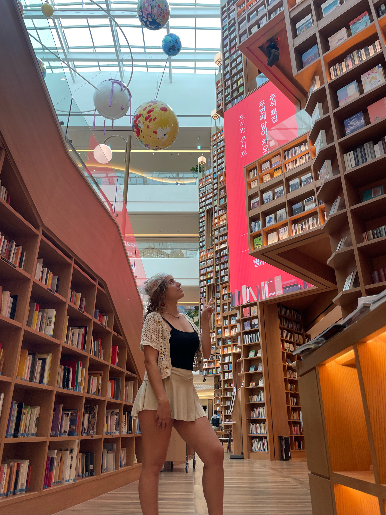 In a bright, open-air indoor space, likely a bookstore or library within the Starfield Library in Suwon-si, South Korea, a young woman stands on a wooden floor, looking up. She wears a black tank top, a light-colored crochet cardigan, and a beige skirt. A patterned headscarf adorns her curly hair, and she wears necklaces and earrings. Her right hand is raised, and her left hand is extended slightly to her side. The background features towering wooden bookshelves filled with books, stretching upwards and disappearing out of frame. Above, a large red digital display with Korean text is visible between shelves. Several large, spherical, colorful art installations are suspended from the ceiling. The architecture of the space includes glass railings and a large, open ceiling with visible beams and skylights, suggesting a modern, multi-level structure. The overall ambiance is one of contemporary design and abundant knowledge.