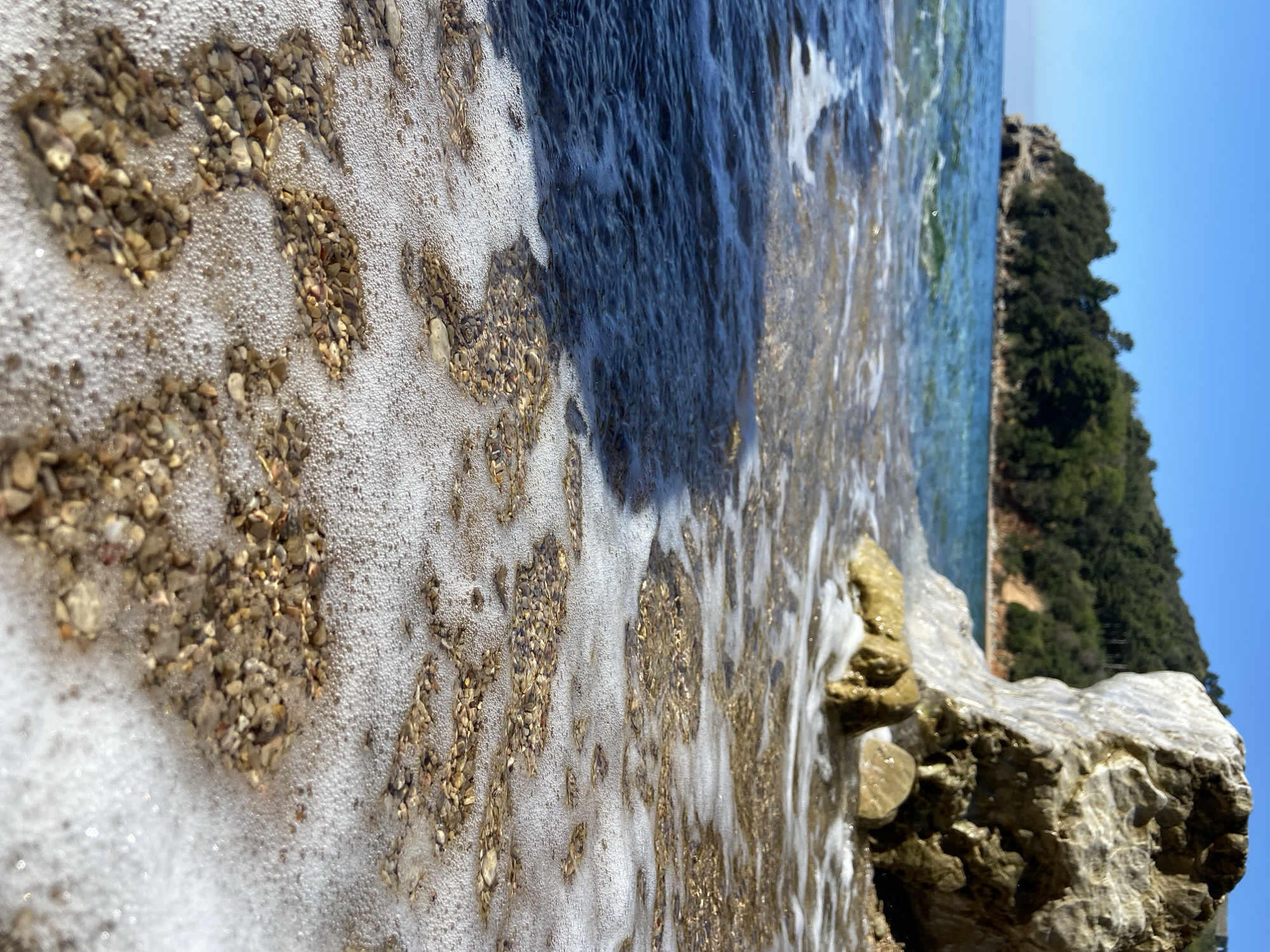 The image depicts a close-up view of the shoreline at a beach, possibly in or near Budva, Montenegro. The beach is characterized by small pebbles and stones being washed by the foamy surf. The water transitions from clear to darker hues as it deepens. In the background, a rocky coastline covered with vegetation rises against a clear blue sky. Large rock formations are visible in the foreground. The scene is observed from a low vantage point, emphasizing the textures of the sand, water, and rocks.