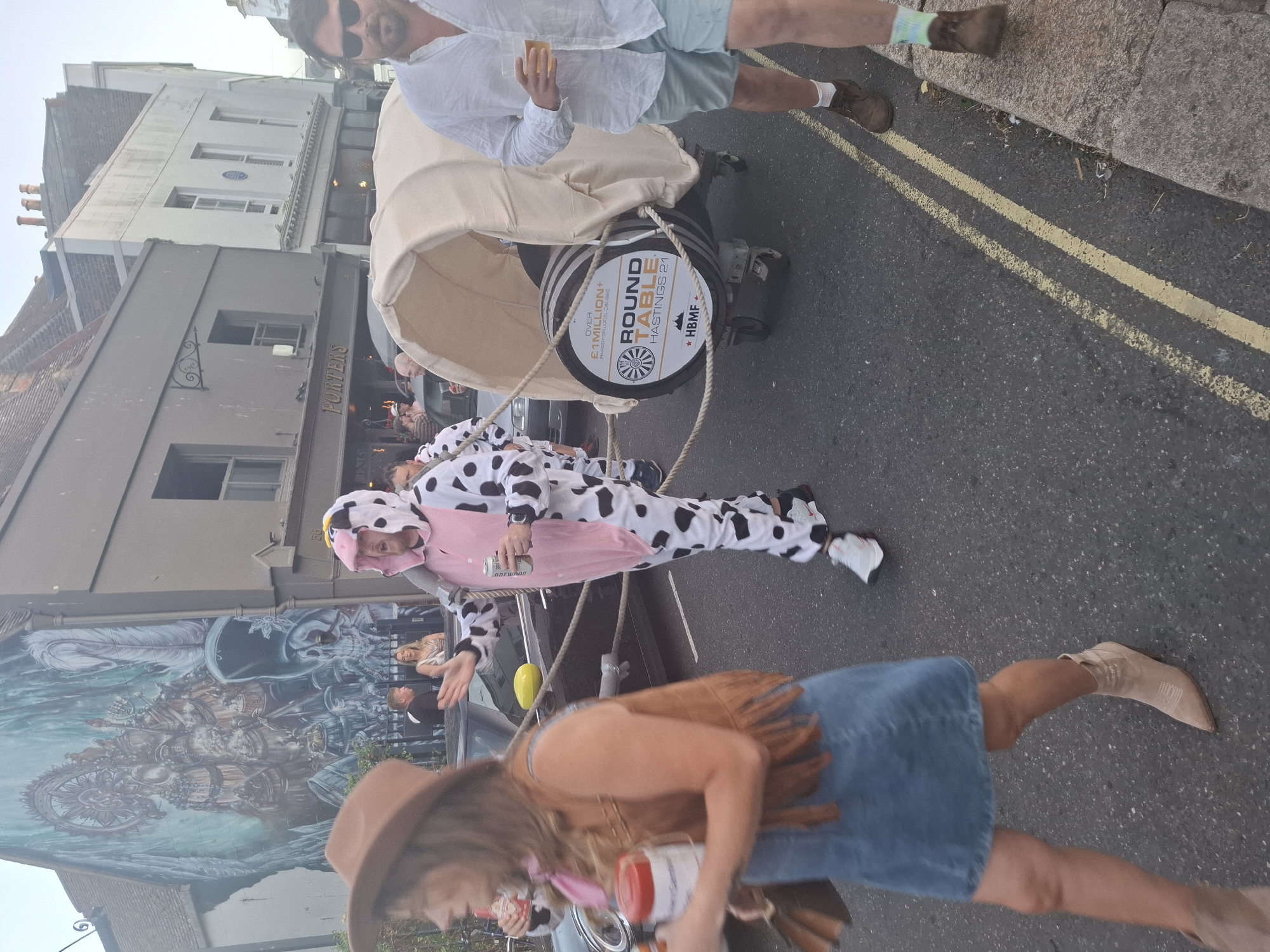 In St Leonards, United Kingdom, on a street with buildings and a mural, a person in a cow costume is pulling a barrel on a cart. The barrel has a logo that reads "ROUND TABLE HASTINGS 21" and "OVER £1MILLION+ RAISED FOR LOCAL CAUSES". Another person in a cowboy hat and vest is walking in front of the cow, holding a red and white container. A man in a white shirt and blue shorts is walking on the right side of the street, holding a drink. The scene appears to be a street event or parade.