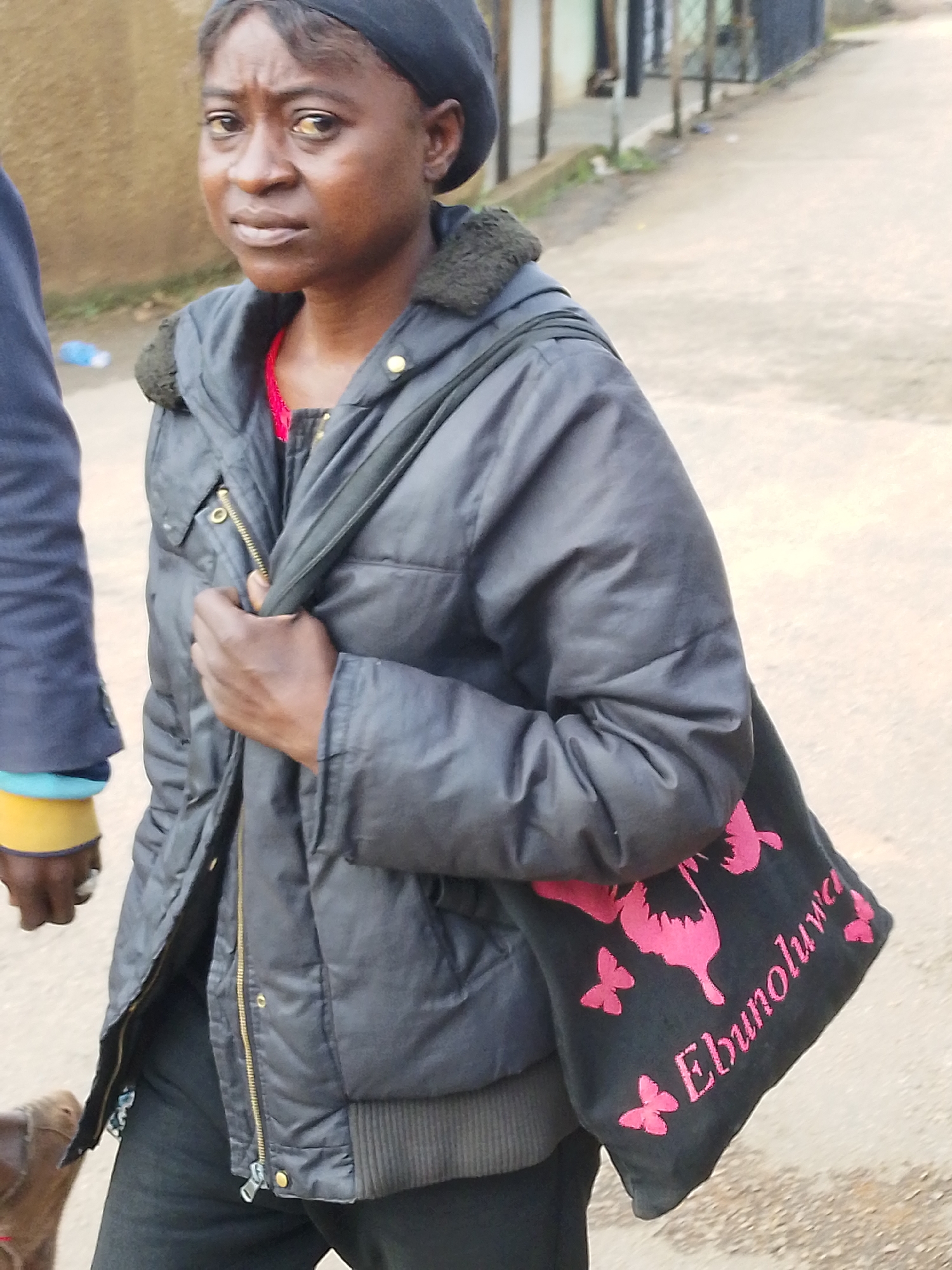 A full, waist-up shot shows a Black woman from the shoulders up, walking outdoors on a paved surface. She is wearing a black, quilted, hooded jacket over a red top. The jacket is zipped open, revealing a hint of the red top. A black crossbody bag with pink embroidered roosters and butterflies, and the word "Ebunoluva" in pink script, is slung across her chest. Her left hand rests on the strap of the bag. Her right hand is partially visible, holding the bag. She is wearing a black beret. Her facial expression is somber, with her eyes looking to her left, and her mouth slightly open. Her skin is dark, and her eyes are brown. She has a scar above her left eyebrow and a slight dark discoloration under her right eye. Her ears are small and close to her head. The background consists of a blurred, outdoor environment with a beige wall on the left and a fence and buildings in the distance on the right. There is a white and blue plastic bottle on the ground to the left of the woman. The lighting appears to be natural daylight.
