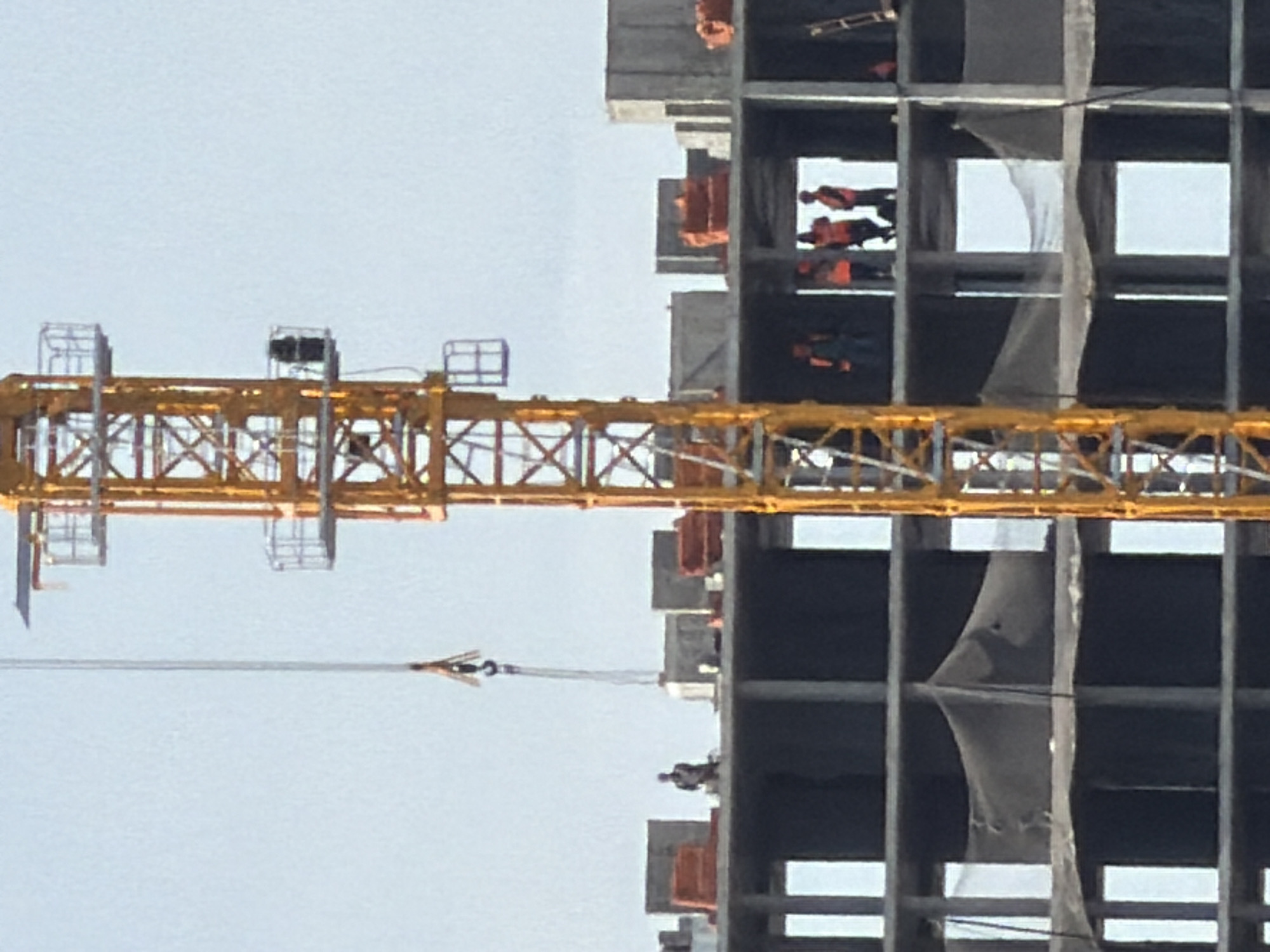 The image depicts an active construction site in Chaikovsky, Russia, under a clear, bright sky. A prominent yellow-orange latticework tower crane occupies the right side of the frame, displaying multiple platforms and a visible operator's cab on its mast. A cable with a hook assembly descends from the crane's upper structure, centrally located above the building.
Adjacent to the crane, a multi-story reinforced concrete building is under construction, presenting exposed concrete floor slabs and vertical columns that define numerous open apertures. On the uppermost completed floor, one individual, dressed in dark attire, is positioned near concrete barriers. On the floor directly below, a group of four construction workers, uniformly wearing high-visibility orange safety vests and hard hats, stand within an open structural bay. One worker in this group appears to wear a dark overall beneath the vest. On the floor below that, a single worker in similar attire is visible on the far left of an open bay.
Orange construction forms or protective barriers are distributed across the upper concrete surfaces of the building. Dark protective netting is visibly stretched across multiple openings on the lower visible floors of the structure. A metallic ladder rests against the building's exterior on the far right side of the frame.