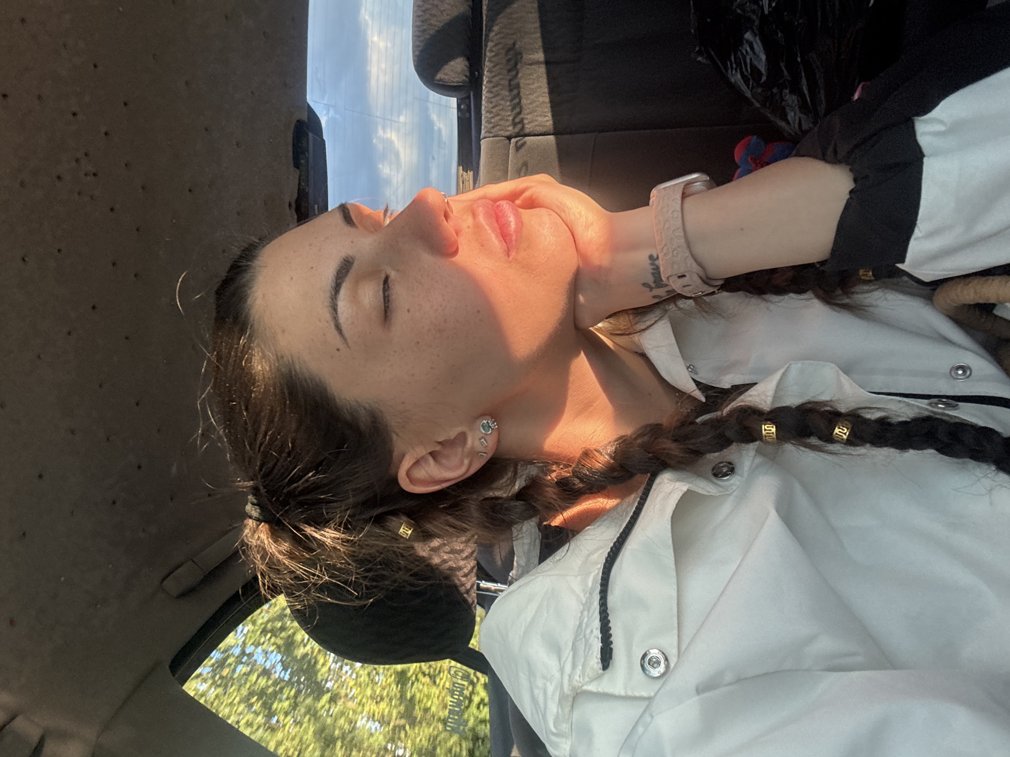 The image shows a young woman with her eyes closed, resting her head on her hand while sitting in the back seat of a car. Sunlight illuminates her face, creating a warm glow and casting shadows. She has her hair in two braids, adorned with gold hair accessories. She is wearing a white jacket and a pink watch. In the background, a black car seat is visible, and outside the car window, trees and a cloudy sky can be seen. The setting appears to be a rural area, possibly the outskirts of Shiroki dol, Bulgaria, given the natural environment and the sunlight characteristic of late afternoon.