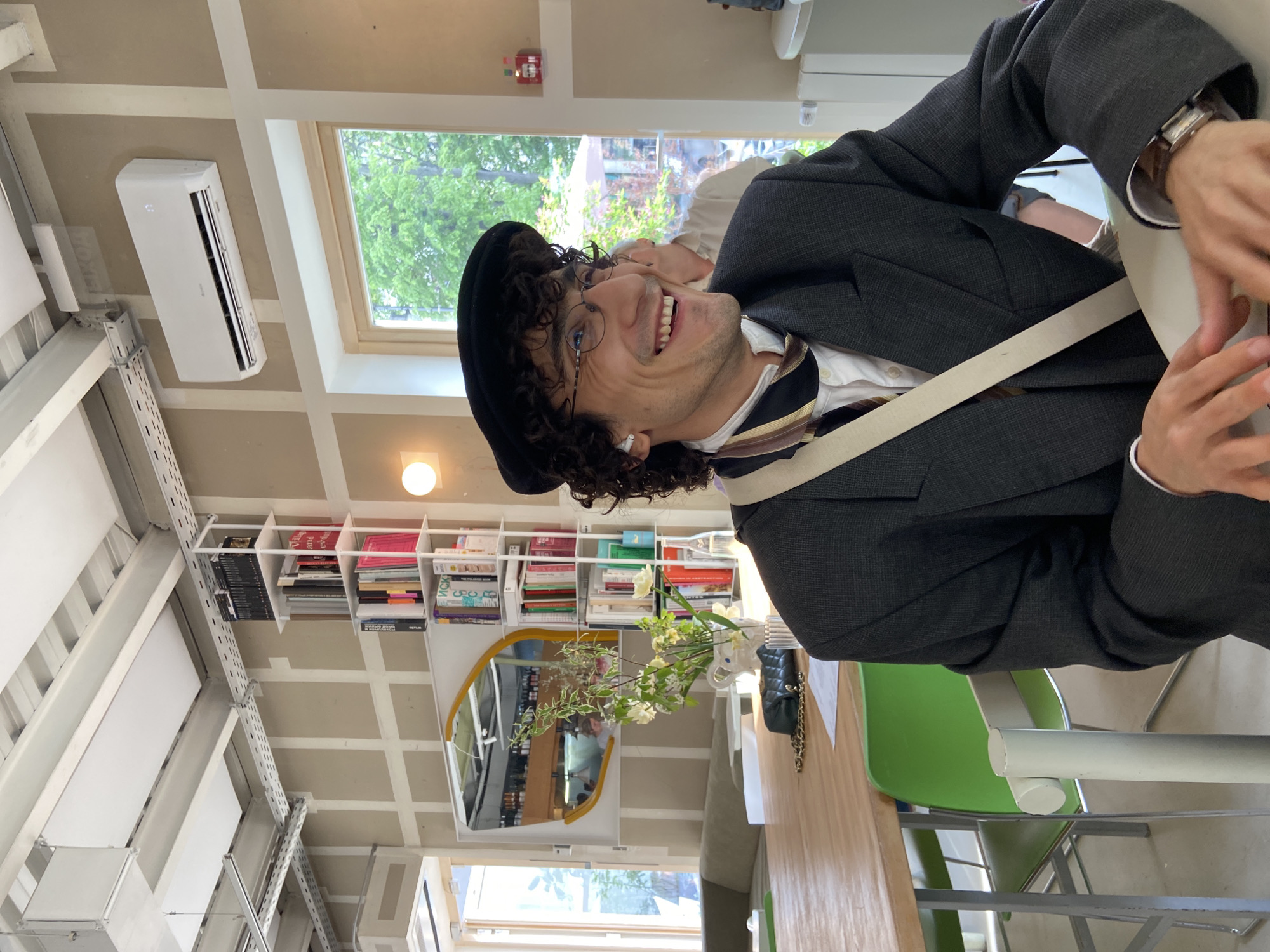 A man with curly hair, wearing a black beret and glasses, is seated in what appears to be a cafe or restaurant. He's dressed in a dark grey suit, white shirt, and striped tie, and has a light-colored strap across his chest. The environment includes an air conditioning unit mounted on the wall, shelves filled with books, a mirror reflecting the interior, a vase of flowers, and a green chair. The presence of books and unique decor suggests a cafe with an intellectual or artistic ambiance. Given the style of the setting, the image may depict a cafe in a cosmopolitan city, but without further context, it's not possible to determine if it is in Moscow, Russia.