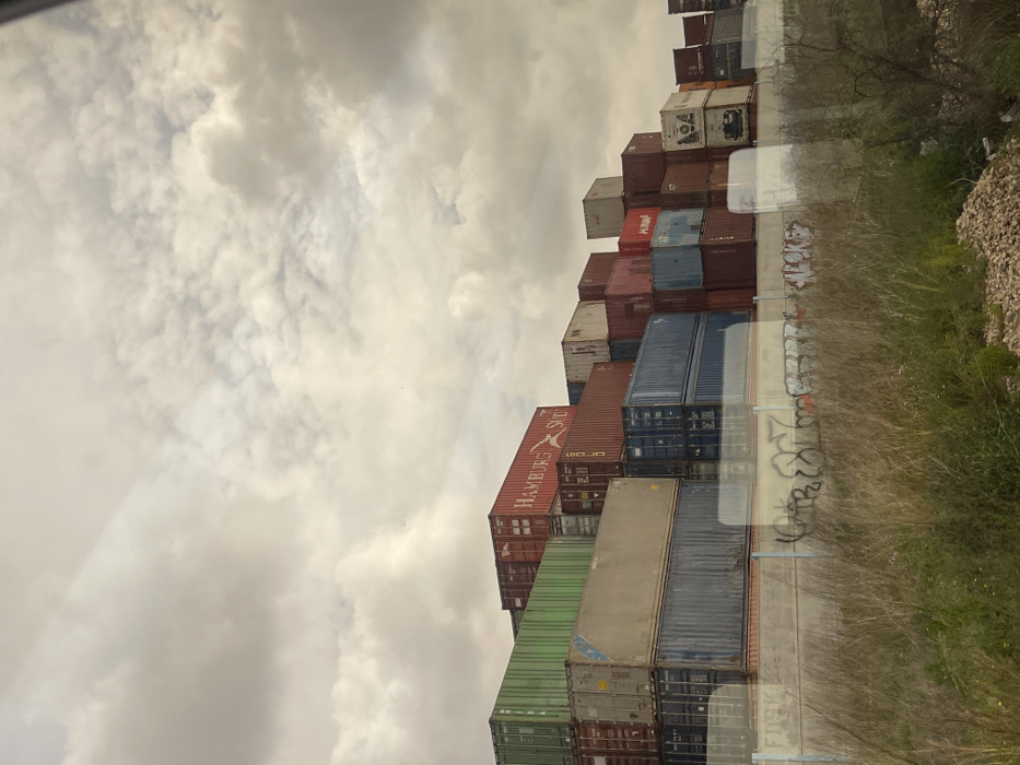 The scene depicts a large-scale storage facility for intermodal shipping containers in Quart de Poblet, Spain, under an overcast sky. Numerous containers of various colors, including green, red, blue, brown, and white, are stacked irregularly, forming a multi-tiered structure extending horizontally across the mid-ground and reaching several stories high. A prominent red container on the left bears the "HAMBURG SÜD" logo. A concrete retaining wall, marked with black and white graffiti, runs across the lower mid-ground. The foreground consists of dry, unmanaged vegetation, primarily tall grasses and weeds, with a patch of gravel and sparse, bare branches visible on the right. Faint reflections are visible across the lower portion of the image, consistent with a photograph taken through a glass pane. No human subjects or active operations are present in the frame.