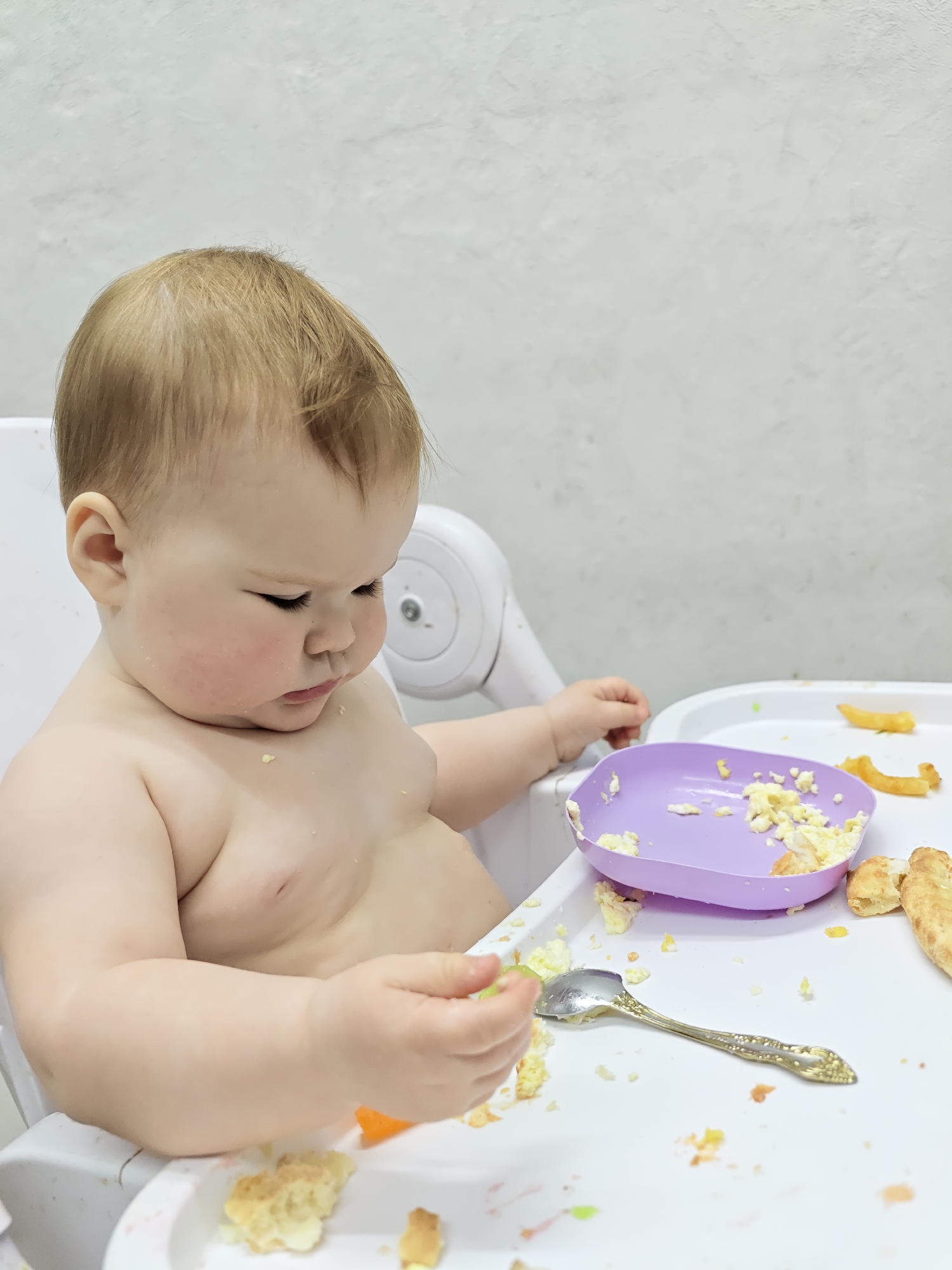 A baby is sitting in a white high chair, looking down at food on the chair's tray. The baby has light-colored hair and is not wearing clothes. On the tray, there is a purple plate containing scrambled eggs, a silver spoon, and other food scraps. The background is a light gray wall. There is no indication of the location being Chaikovsky, Russia.