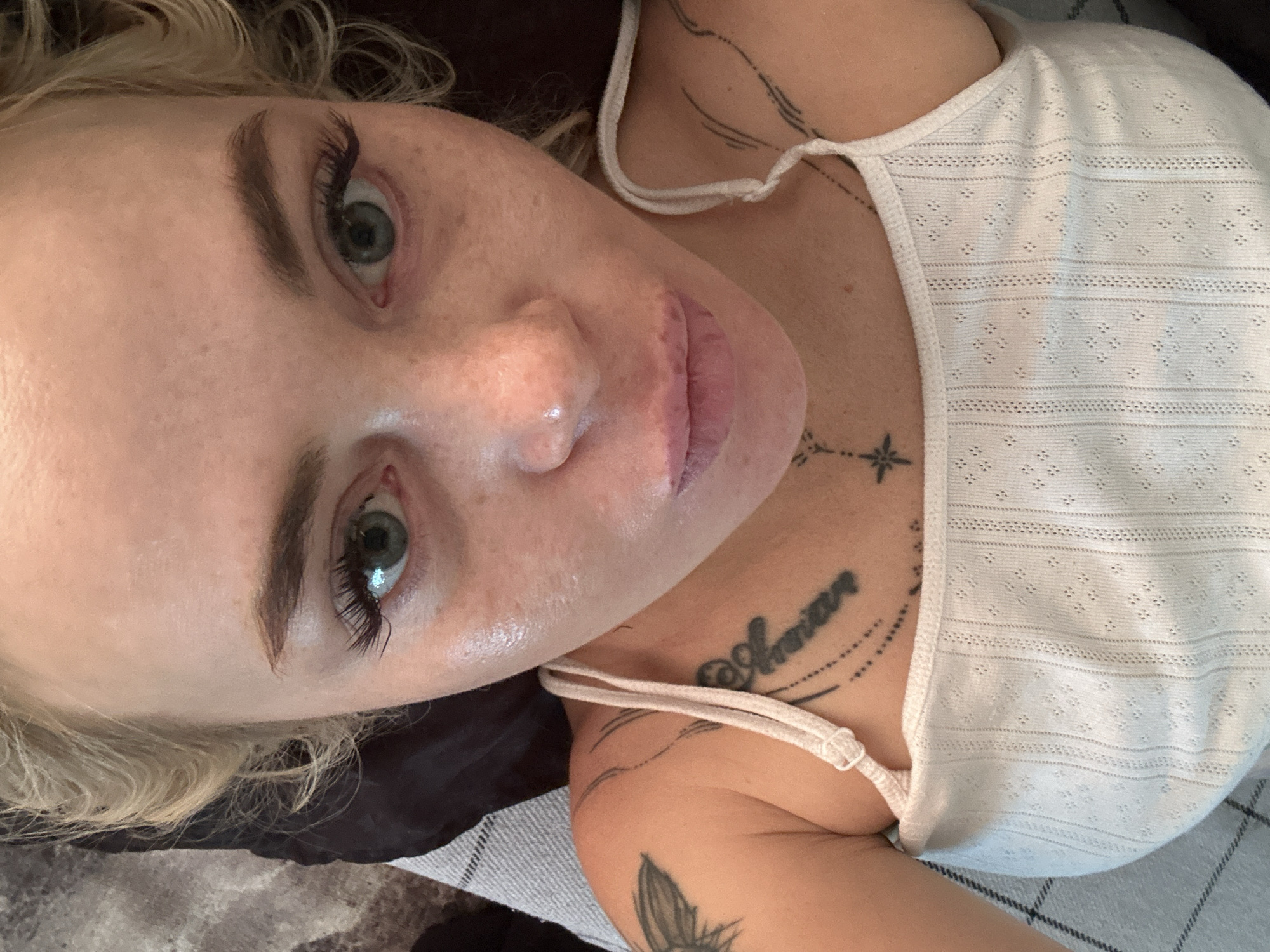 A close-up selfie captures a young Caucasian woman with blonde hair and green eyes. She is lying down, wearing a white tank top. Tattoos are visible on her left shoulder and chest. The background includes a dark pillow and a grey and white checkered blanket. The lighting is bright, highlighting her facial features, including freckles and makeup.