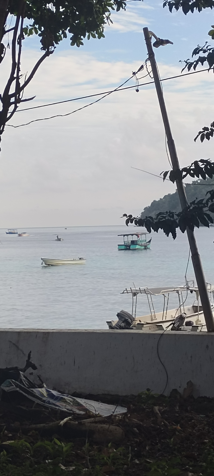 The image shows a coastal scene in Sabang, Indonesia, with a body of water in the foreground and background. Several boats are visible on the water, including a small white boat in the middle ground and a larger boat with a green hull further in the distance. In the foreground, there is a concrete barrier, and beyond it, an area with debris, including branches and what appears to be a tarp. Overhead, there are power lines and foliage from trees. The sky is partly cloudy. The scene appears to be a daytime view of a harbor or bay area.