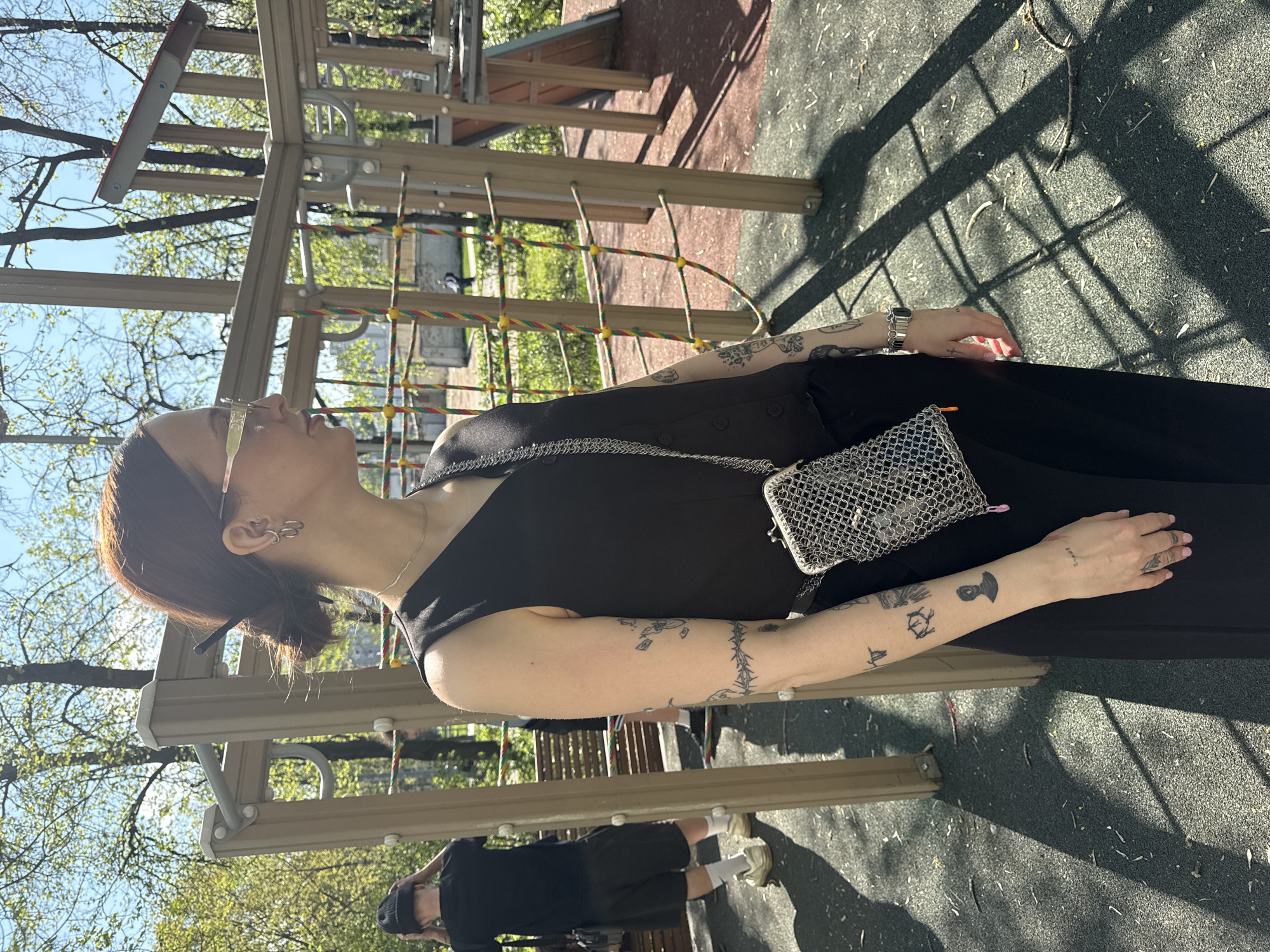 A woman is standing in a playground in Moscow, Russia. She is wearing a black dress, sunglasses, and a silver purse. Her arms are covered in tattoos. In the background, there is a playground structure made of wood and rope. A person wearing black shorts, black t-shirt, white sneakers and white socks is in the left foreground. The ground is covered in a gray material with dark spots. The trees are visible in the background. It is daytime and sunny.