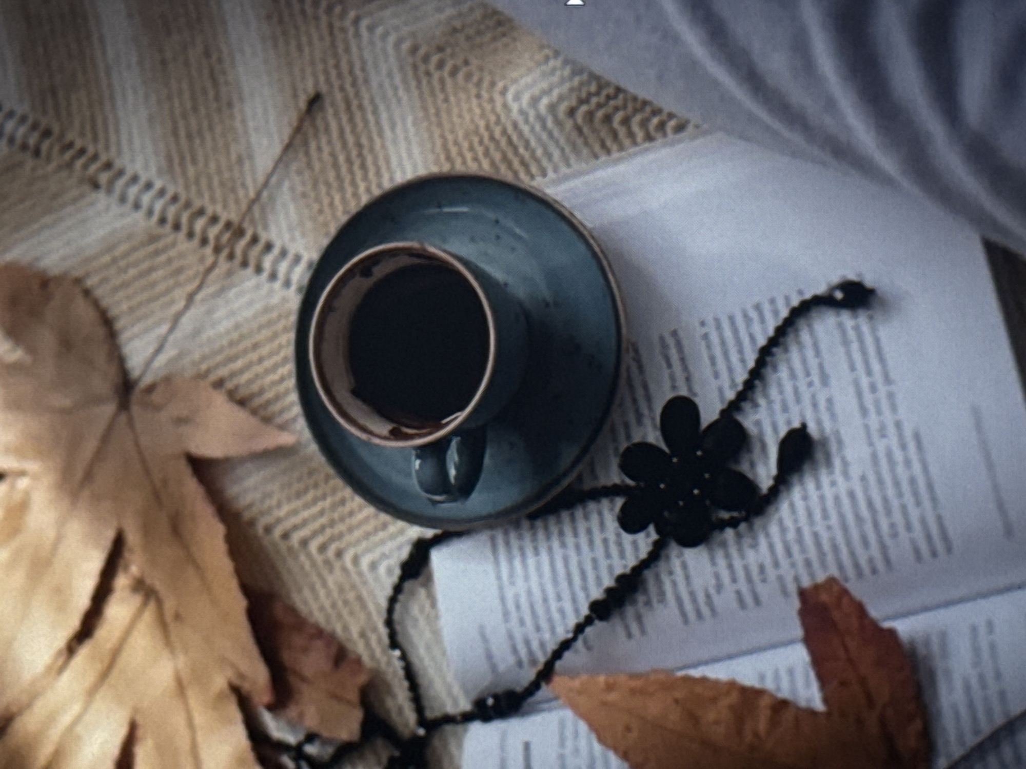 A flat lay composition features a coffee cup on a saucer, placed on an open book. The book lies on a textured, light-colored blanket with a woven pattern. To the left of the cup, a dried, light brown leaf is partially visible, and to the lower right, another similar leaf rests on the book. A black beaded necklace with a black floral charm is draped across the book and around the coffee cup. The coffee in the cup appears dark. The overall lighting suggests a soft, natural light, possibly indicating daytime, but the exact time is not discernible. The scene evokes a sense of quiet contemplation or relaxation. There is no visible text in the image. The context provided places this scene in Eyüpsultan, Turkey, suggesting a domestic or cozy setting within that location.