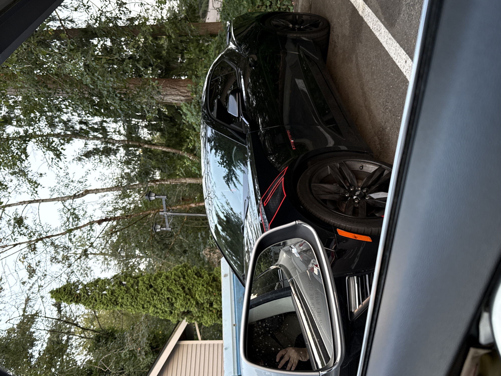 The image, taken in Gig Harbor, United States of America, shows a black Chevrolet Camaro parked outdoors. The car has red racing stripes on its front fender and hood. In the foreground, a side-view mirror reflects a person's hand holding a phone, possibly taking the picture. The background features trees and a building.