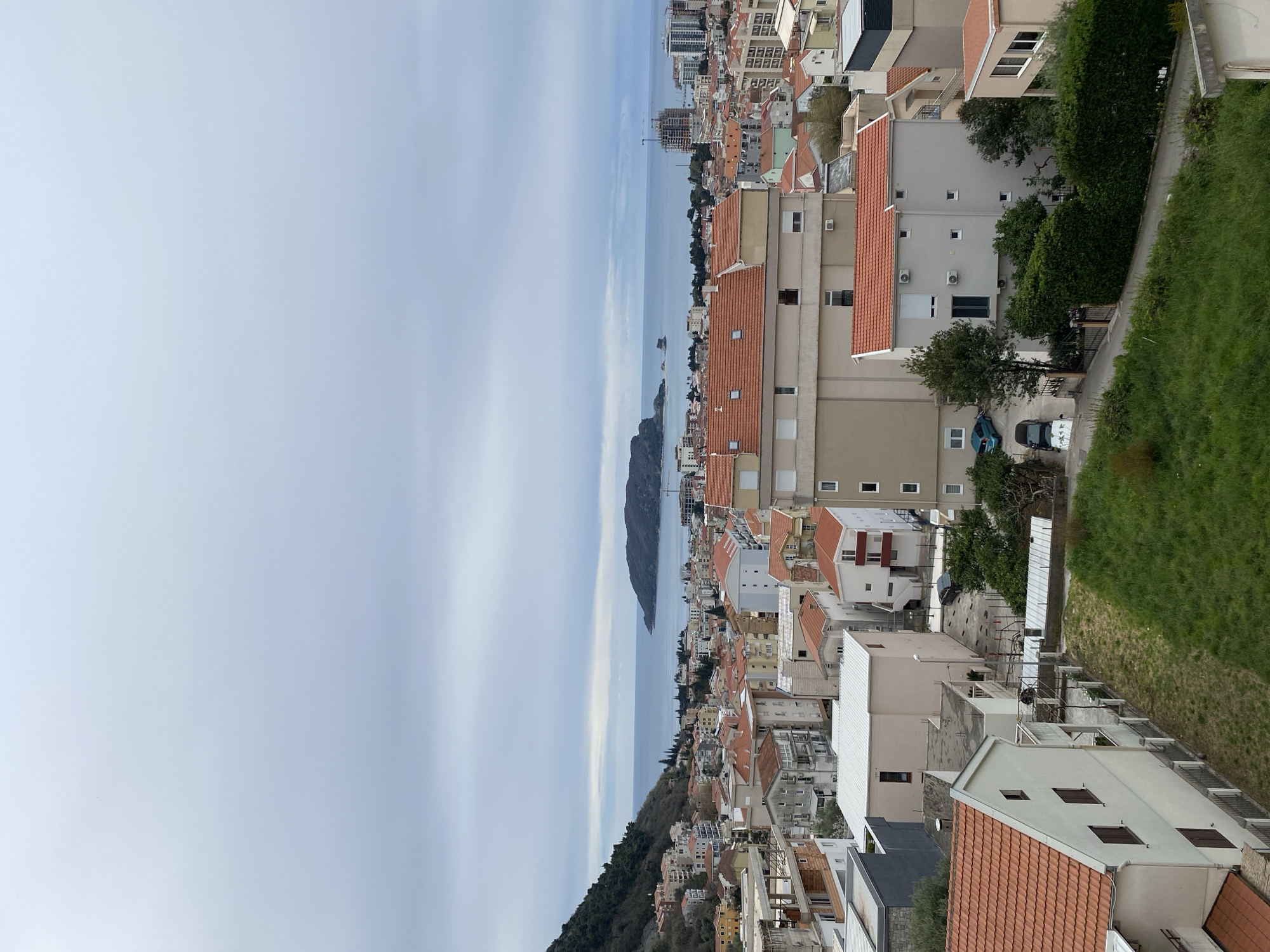 An aerial view of Budva, Montenegro, showcases a coastal cityscape dominated by buildings with predominantly red-tiled roofs. The Adriatic Sea extends from the city towards a mountainous island in the distance. The foreground features residential buildings and a green hillside. Cars are parked in the driveways below. The sky is overcast, casting a diffused light over the scene.