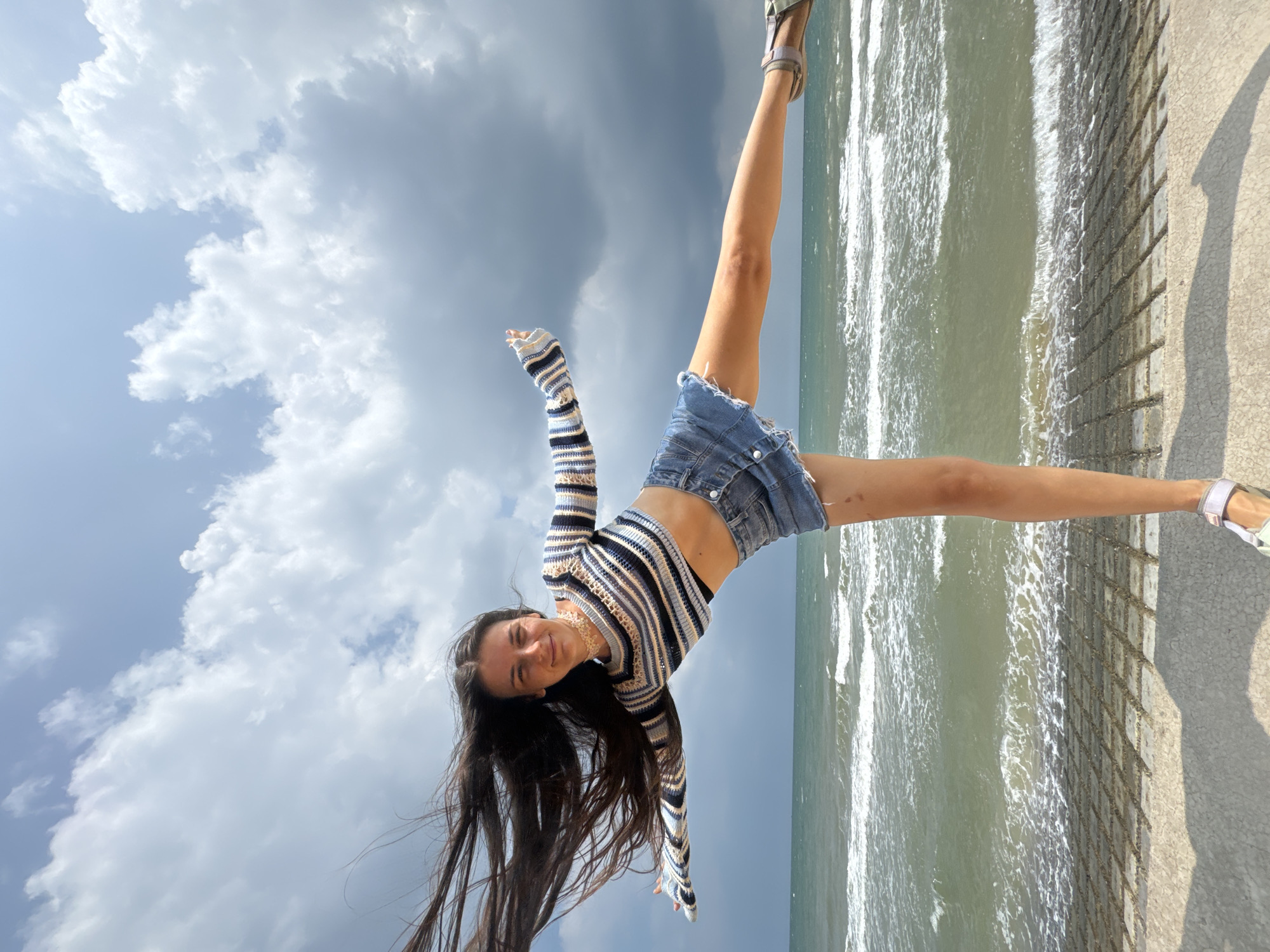 A young woman with long, dark hair is captured in a dynamic pose on a paved walkway near the sea. She is wearing a striped, cropped sweater and denim shorts. Her right leg is extended forward, and her arms are outstretched. The sea is visible behind her, with waves breaking on the shore. The sky is filled with dramatic, grey clouds. The location is identified as Phu Thuy Ward, Vietnam.