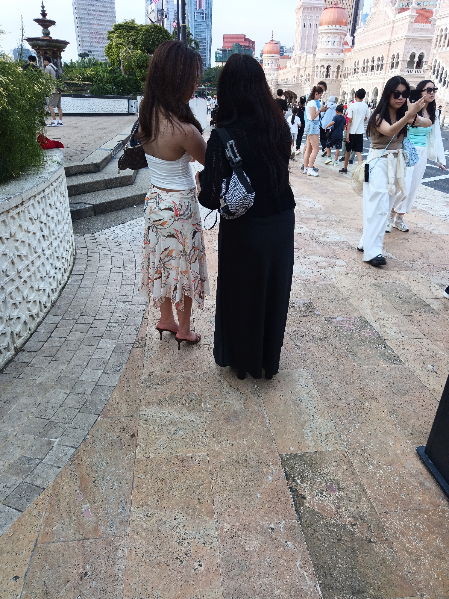 This image depicts two women standing on a paved walkway in front of a historical building in Kuala Lumpur, Malaysia. The building, likely the Sultan Abdul Samad Building, features distinct Mughal-inspired architecture with domes and arches, illuminated by daylight. The walkway is composed of large, light-colored stone tiles with some discoloration.
The woman on the left is wearing a white strapless top and a floral-patterned skirt with a high slit. She has dark hair and is wearing heeled sandals. She is holding a dark-colored handbag. The woman on the right is wearing a black long skirt and a black top. She has long dark hair and is wearing black shoes. A patterned shoulder bag is visible on her right shoulder.
In the background, other people are walking, some of whom are taking photographs. The environment appears to be a public space, possibly a tourist attraction, with some greenery and modern buildings visible in the distance. The overall lighting suggests it is daytime, with a slightly overcast sky.