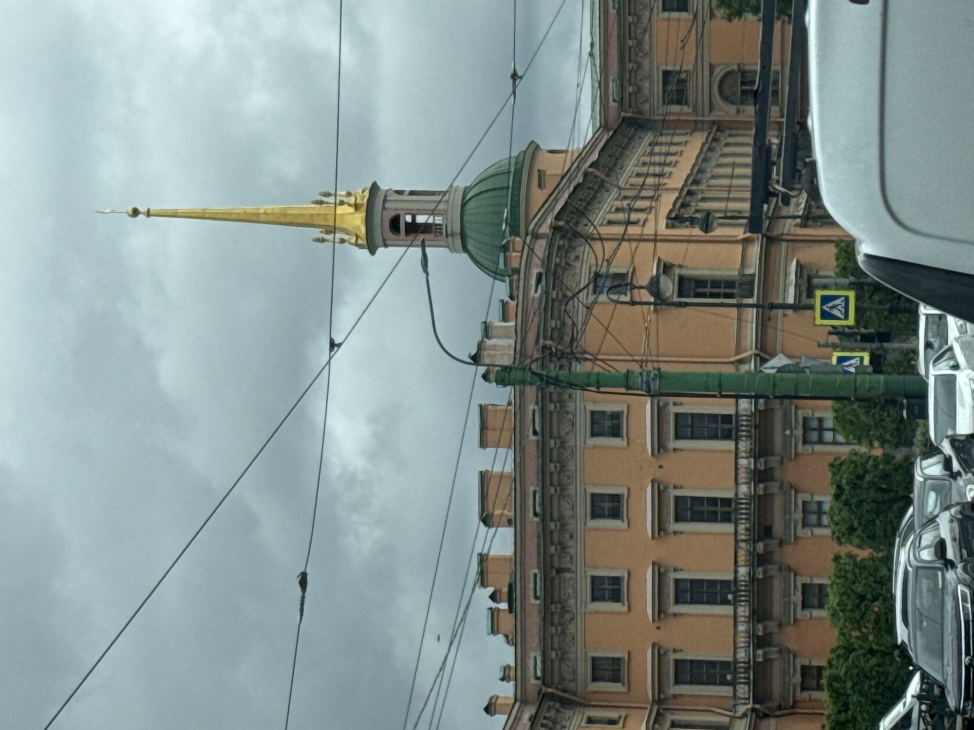 This image, likely taken in Saint Petersburg, Russia, presents a street view dominated by an orange-toned building with ornate window detailing and a distinctive green dome topped with a golden spire. The spire extends into a cloudy sky. Power lines crisscross the view. A pedestrian crosswalk sign is visible on a green pole. Parked cars fill the right side of the frame.