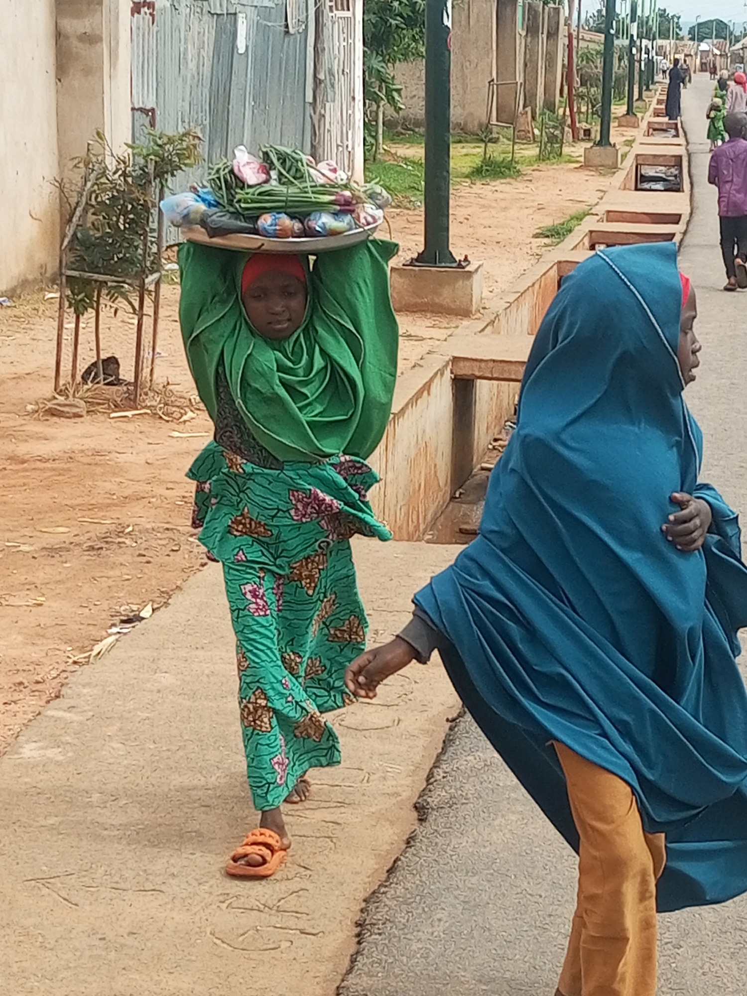In Tempure, Nigeria, a young girl wearing a green patterned dress and an orange headscarf walks along a dirt path. She carries a metal tray laden with produce, including green beans and red onions, balanced on her head. To her right, another young person, also wearing a blue headscarf and brown pants, walks alongside her, their hand resting on her arm. In the background, other individuals are visible walking along a paved street lined with streetlights and concrete dividers. The scene depicts a typical street view in a Nigerian city.