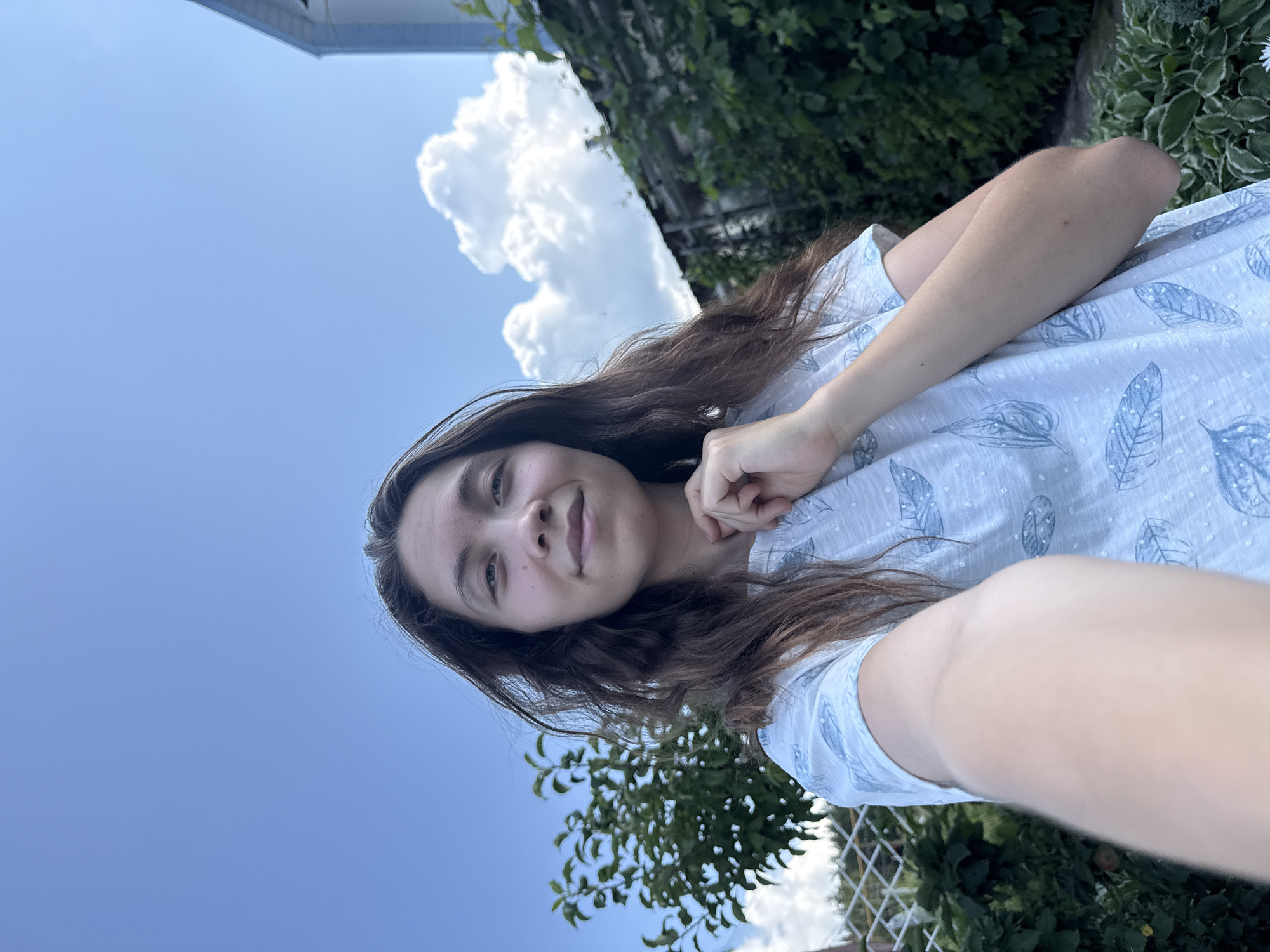 A young woman with long, dark, wavy hair is taking a selfie outdoors. She is wearing a light-colored t-shirt with a blue feather pattern. Her left arm is extended towards the camera, holding it. She is smiling faintly and touching her chin with her right hand. The background shows a clear blue sky with scattered white clouds, green foliage, and a weathered wooden structure. A portion of a white building is visible in the upper right corner. The setting appears to be a rural or suburban backyard. The image was captured in Arzyanka, Russia.