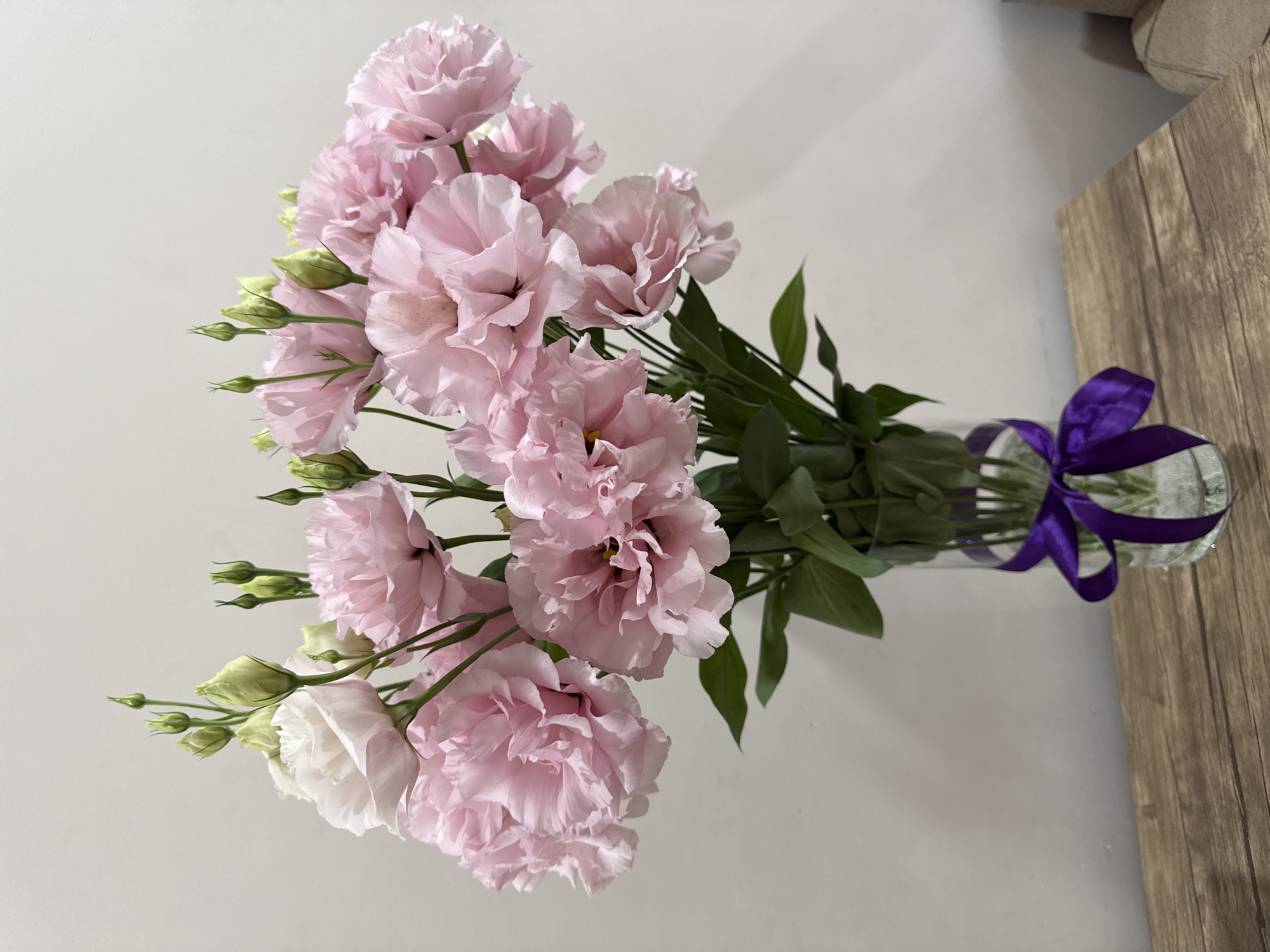 A bouquet of pink and white lisianthus flowers with green buds is arranged in a clear glass vase on a wooden surface. The vase is adorned with a purple ribbon tied in a bow. The background is a plain, light-colored wall. This image does not provide any information about the city of Yerevan, Armenia.