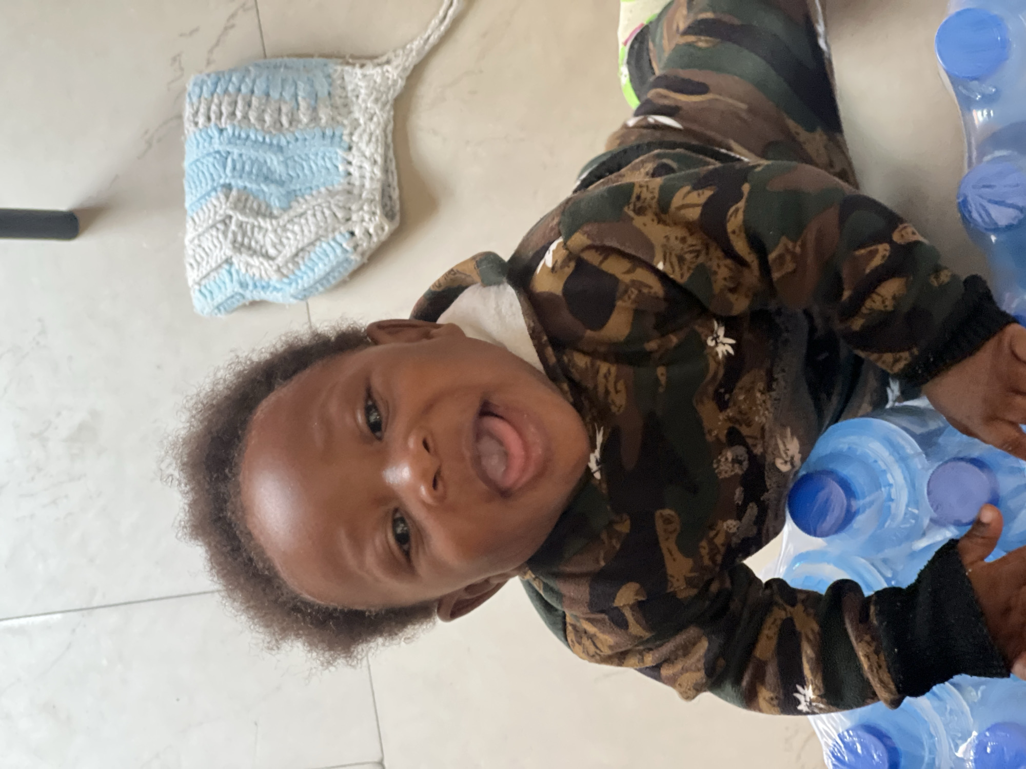 A joyful young child with dark, curly hair and dark skin is captured in a close-up, eye-level shot, lying on a light-colored, possibly tiled floor in an indoor setting in Jos, Nigeria. The child wears a camouflage-patterned hoodie or top, featuring shades of brown, green, and black, with small white designs. Looking directly at the camera, the child has a wide-open mouth with a visible tongue, conveying a playful scream, laughter, or an excited expression.
To the child's upper left, a light blue and white crocheted item with a chevron pattern is visible on the floor. To the child's right, a stack of clear plastic water bottles with blue caps, still in their transparent plastic packaging, is present, with one of the child's hands resting near them. The bright, even lighting suggests a daytime scene. The overall mood is candid and full of childlike happiness. No visible text is present in the image.