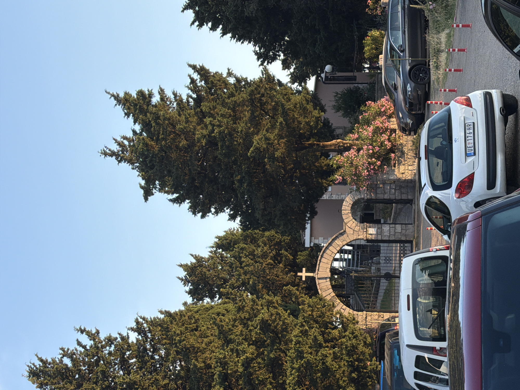 This media file depicts an outdoor scene with several cars parked along a street. In the foreground, a white Peugeot 207 is visible, its license plate reading "UE 176 HR". Next to it, a dark-colored SUV is parked. Further down the street, another dark-colored car is parked. A white van is also present in the scene.
The background features a building with an arched entrance, made of stone. A small cross is visible on the building. Tall trees with dense foliage surround the building and the street, casting shadows. Pink flowers are blooming on a bush next to the building.
The image appears to be taken during the daytime, with clear blue skies. The lighting suggests it might be late afternoon or early morning. The scene is static, with no people visible. The presence of parked cars and the general setting suggest a residential or public area. The overall mood is calm and ordinary.