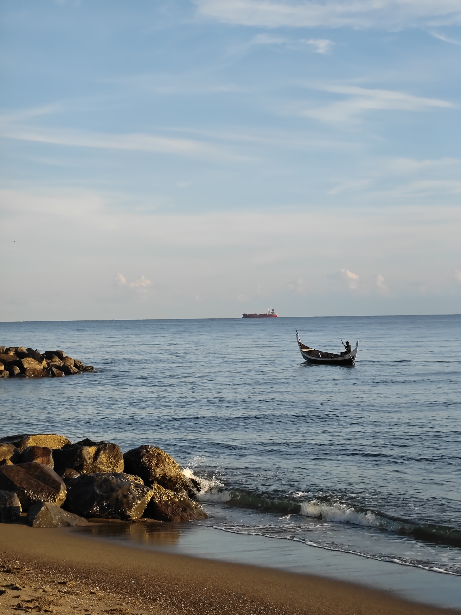 The image shows a coastal scene in Lhokseumawe, Indonesia. A man is rowing a small, traditional boat in the sea. In the background, a large cargo ship is visible on the horizon. A rock barrier extends into the water from the sandy beach in the foreground, where small waves break. The sky is light blue with scattered white clouds.