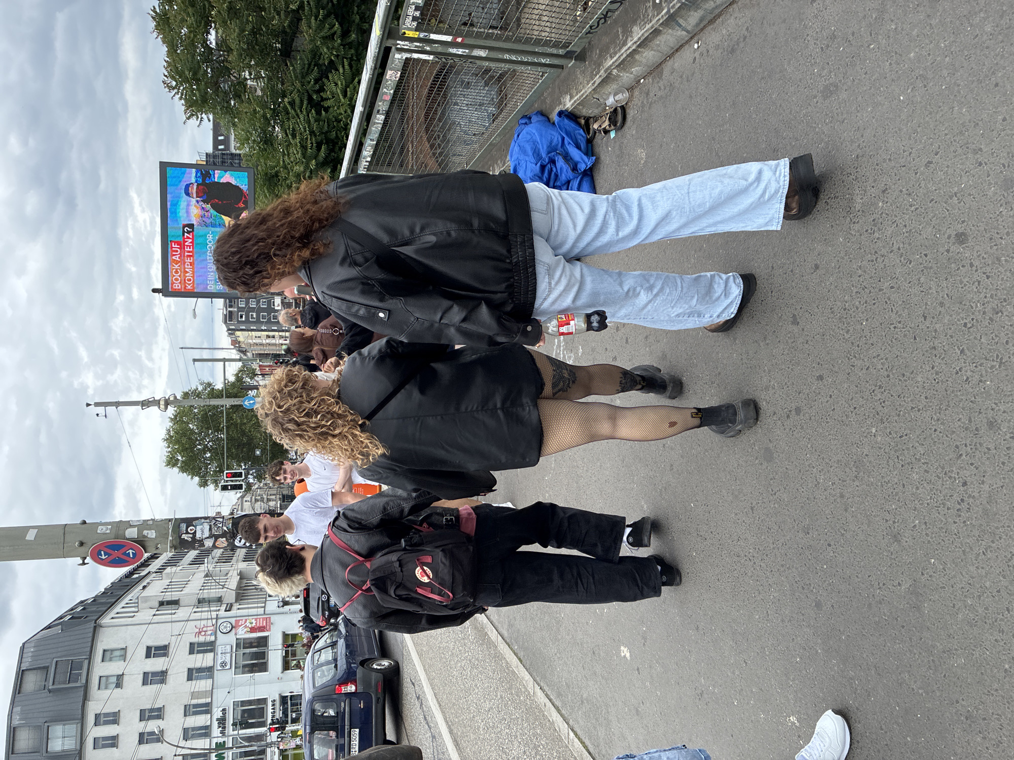 In Berlin, Germany, on a cloudy day, a group of four individuals walks along a paved pedestrian pathway adjacent to a street with vehicular traffic. In the foreground, two individuals with curly hair walk away from the camera, one wearing a black leather jacket, fishnet stockings with a heart detail, and chunky black boots, while the other wears a similar black leather jacket and light blue wide-leg jeans. To their left, another individual with blonde hair and a black backpack also walks away from the camera, dressed in black pants and shoes. Behind them, a young man in a white t-shirt walks towards the camera. In the background, a dark blue van is parked, and buildings with multiple stories line the street. A large digital billboard displays an advertisement with German text and an image of a person engaged in outdoor sports. The pathway is bordered by a metal fence with graffiti. A blue tarp and sandals are visible on the ground near the fence.