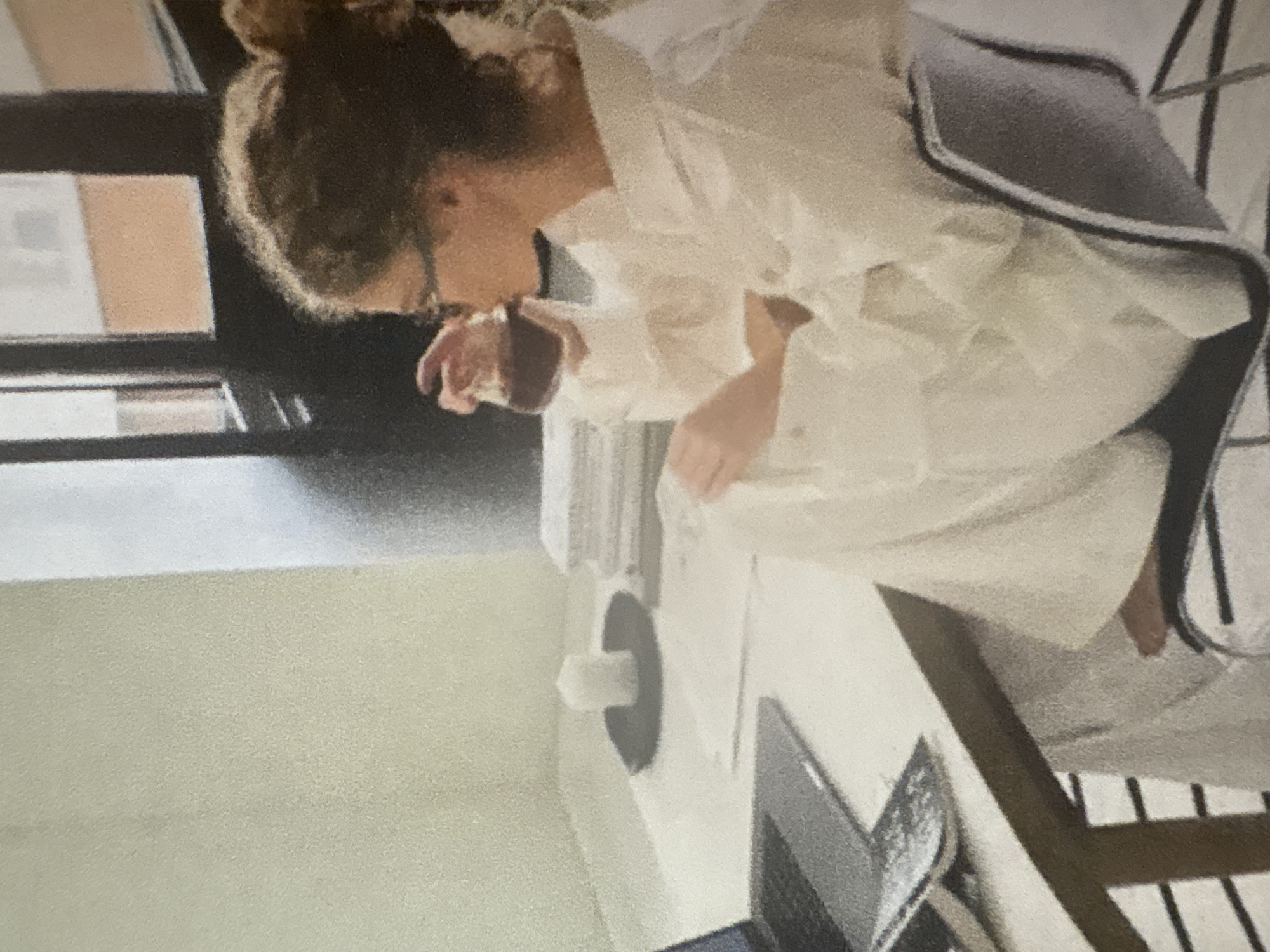 A person with curly brown hair, wearing glasses and a white oversized shirt, is sitting at a desk and drinking from a clear glass. They are seated in a modern chair. On the desk, there is a laptop, papers, a white candle on a dark plate, and a stack of books. The background shows a window with a view of a building. The scene takes place indoors, in what appears to be a home office or a modern living space. There is no indication of this location being in the city of Kazan, Russia.