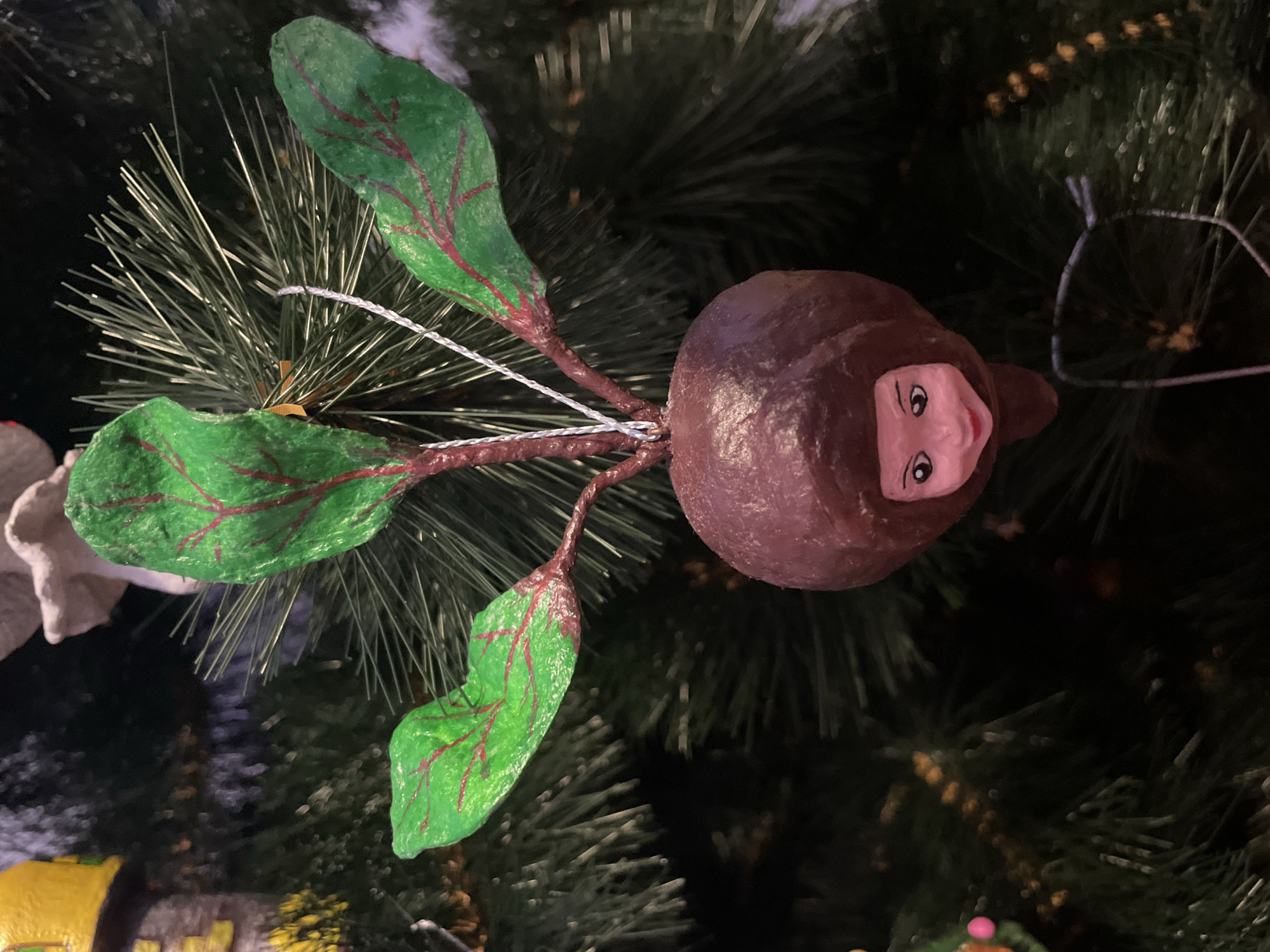 This image features a handmade Christmas tree ornament shaped like a beet. The beet ornament has a small, human-like face with painted eyes and a smiling mouth. It has three green leaves with red veins, attached to reddish-brown stems. The ornament is hanging on a decorated Christmas tree, which is visible in the background, along with other tree decorations. The scene appears to be indoors, likely in a home, given the context of a Christmas tree. The lighting suggests it is evening or nighttime, as is typical for indoor festive decorations. The ornament's whimsical design, with its anthropomorphic face, adds a playful and charming element to the holiday decor. The background is dark and out of focus, emphasizing the ornament as the main subject. There is no visible text in the image.
