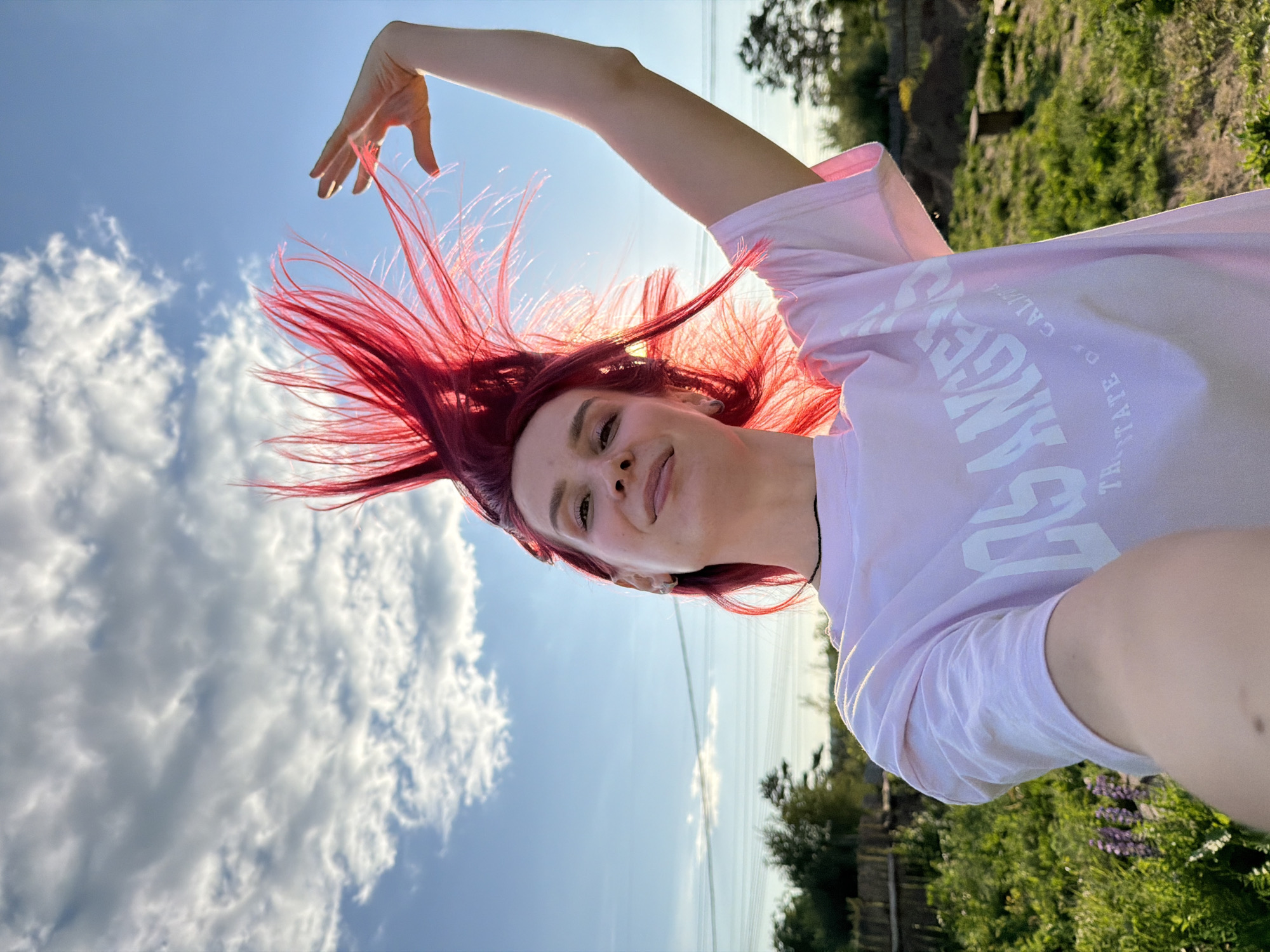 An outdoor selfie depicts a young woman with vivid red hair, styled dramatically upwards as if in motion or wind-blown, against a bright blue sky with scattered white clouds. The sun, positioned behind her, backlights her hair and the right side of her face. She smiles subtly towards the camera, which she is holding. Her left arm is raised above her head, with her hand partially visible against the sky. She wears a light-colored t-shirt bearing the text "LOS ANGELES" and "THE GOLDEN STATE OF CALIFORNIA." The background features a green, grassy landscape with small purple flowers (likely lupines) on the left, a wooden fence, and distant trees. Power lines are faintly visible across the horizon. A wooden bench or stool is present in the far right background. The scene is set outdoors, reportedly in the city of Chaikovsky, Russia.