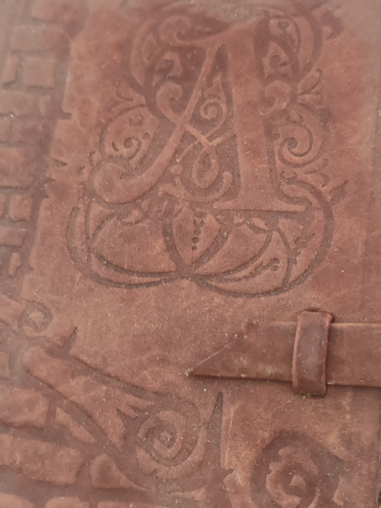 The image shows a close-up of a brown leather surface with embossed designs. The most prominent design is a large, stylized letter "A" with ornate swirls and flourishes. There's a small leather strap with a buckle-like element. To the left, there's a pattern resembling brickwork. The overall texture of the leather appears somewhat rough. There is no information related to the city of Minsk, Belarus.