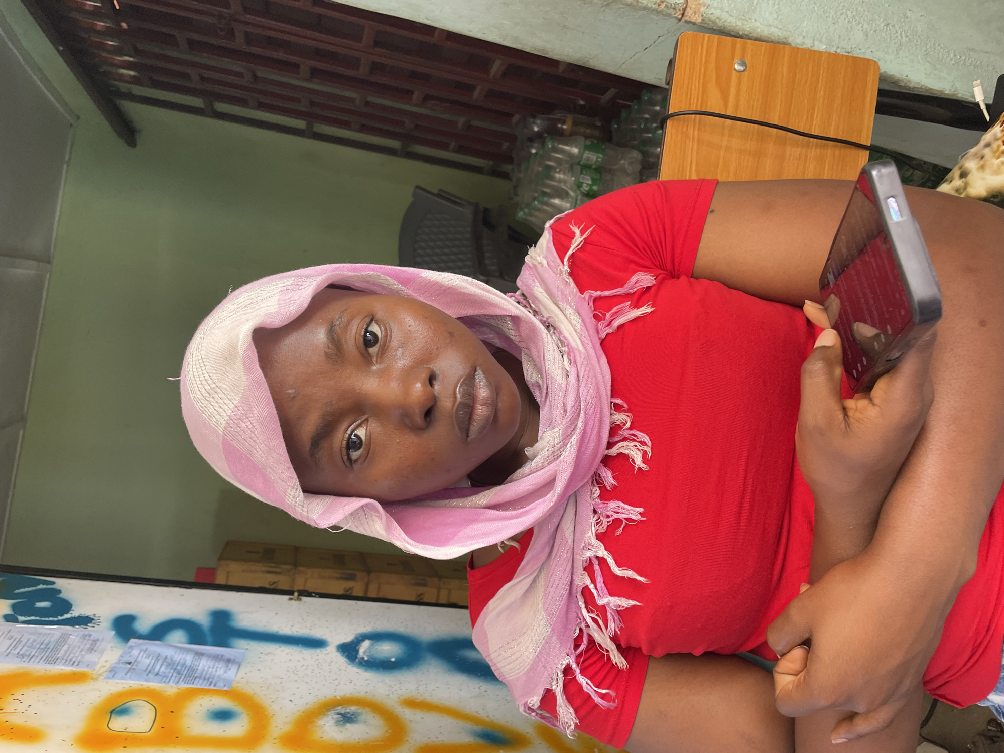 A medium-shot, waist-up, portrait-oriented photograph shows a dark-skinned woman holding a smartphone. The woman wears a red t-shirt and a striped pink and white headscarf. She is looking directly at the camera. Her arms are crossed, and she holds the smartphone in her right hand, angling the screen towards the viewer.
The background includes:
-A green wall with a brown trim.
-Bottled water stacked in the background.
-A brown wooden surface, possibly a desk or table.
-A decorated counter or table, with blue and yellow painted designs and papers on top.
The location is indicated by the subject's appearance and the general environmental conditions suggestive of a residential or business area in the city of Jos, Nigeria. The image is an indoor shot.