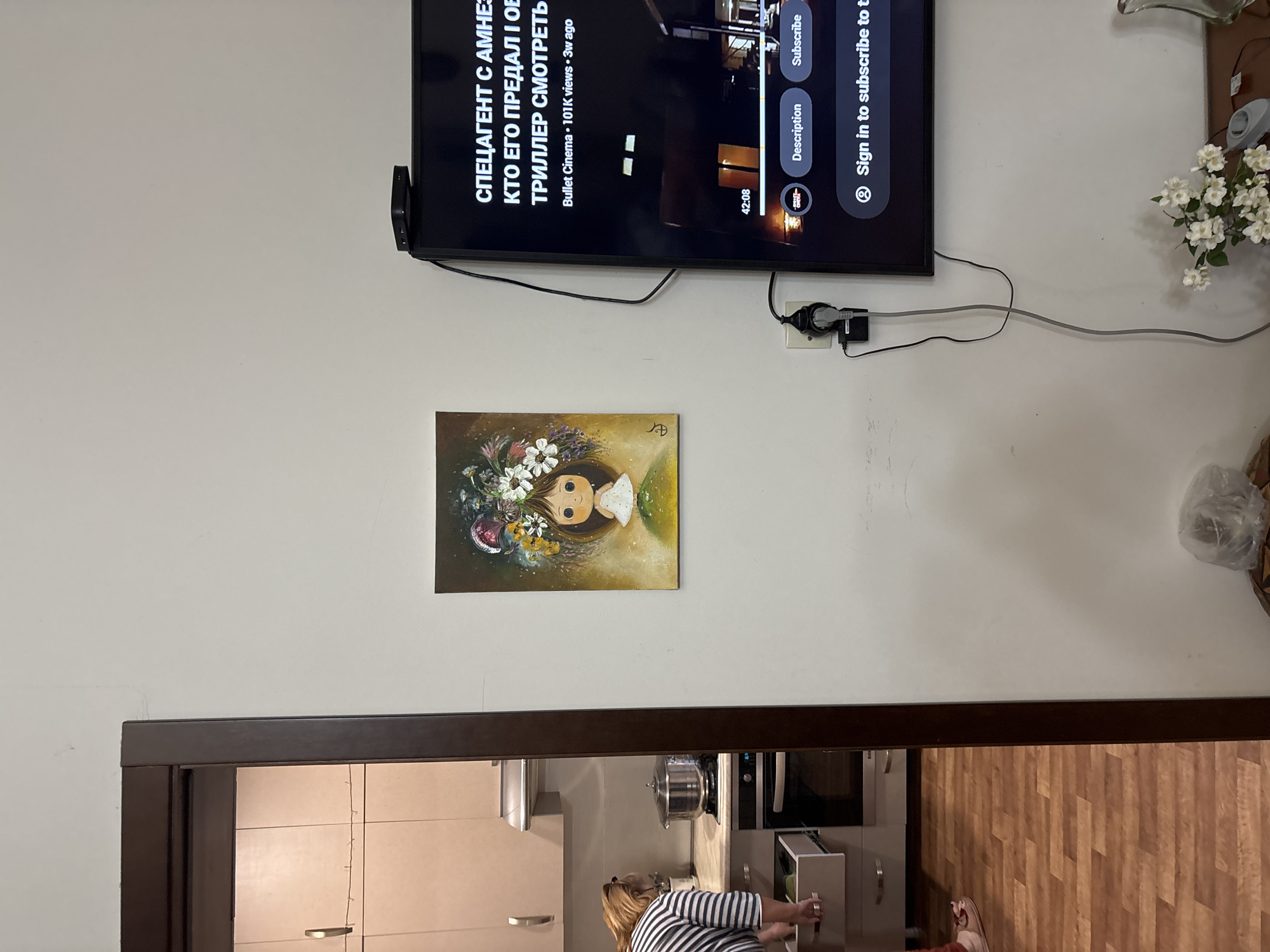 A domestic interior is captured in a vertical orientation, featuring a television mounted on a white wall above a painting of a girl's face surrounded by flowers. Underneath, a doorway leads into a kitchen where a woman in a striped shirt stands at a cabinet. The TV screen displays a video in Russian and has an attached streaming device. Power cords and electrical outlets are visible on the wall. In the upper right, there is a vase with white flowers. The flooring in the kitchen is wood-look planks. The location cannot be determined.
