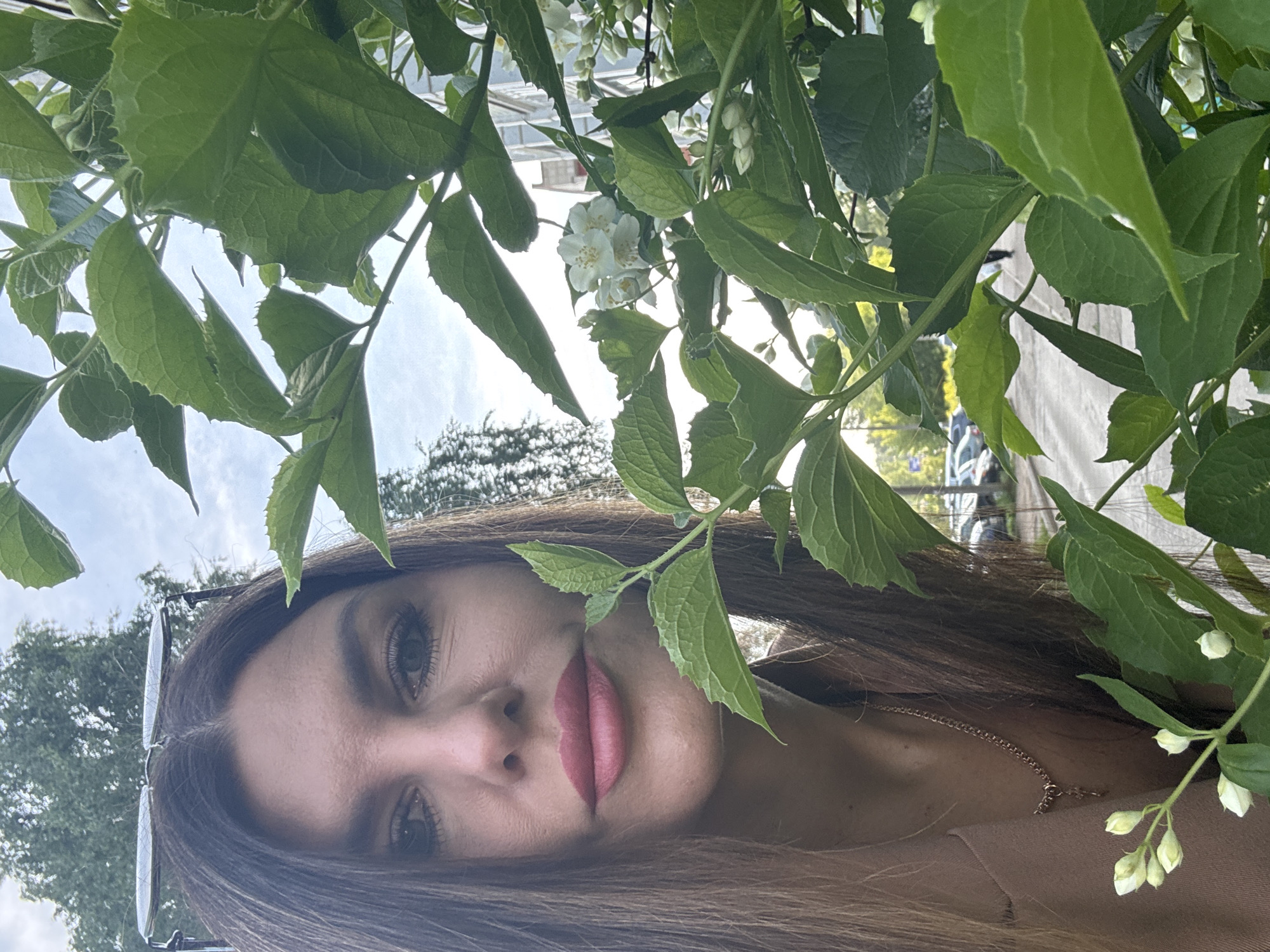 This image is a close-up portrait of a young woman, partially framed and obscured by abundant green foliage and white flowers, taken outdoors in what is stated as Chaikovsky, Russia. The woman, who has long brown hair, dark eyes, and pink lipstick, looks directly at the viewer with a calm, neutral expression. Her face is positioned towards the bottom and right side of the frame (when correctly oriented), with several green leaves and white blossoms overlapping parts of her head and face.
She wears sunglasses pushed up onto her head, and a thin gold chain is visible around her neck, suggesting a light-colored top. The foliage dominating the upper and left portions of the image consists of vibrant green leaves and clusters of small white flowers and buds, characteristic of a flowering shrub like jasmine. These natural elements are in sharp focus, creating a layered depth.
The background, largely out of focus, reveals a bright, overcast or slightly cloudy sky and blurred shapes of distant trees and possibly urban structures. The strong natural light indicates that the photograph was taken during the daytime. The scene conveys a sense of natural beauty and a peaceful, intimate moment captured amidst a lush outdoor setting. There is no visible text in the image.