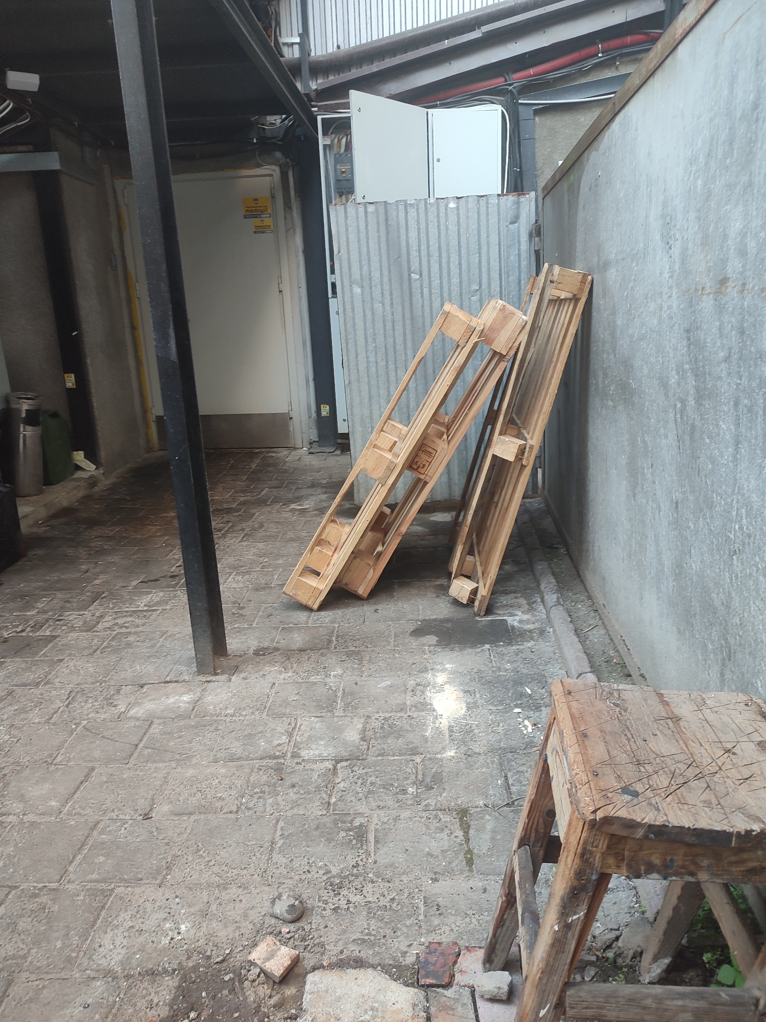 The image shows an outdoor alleyway with a paved floor, likely in Chernivtsi, Ukraine. Two wooden pallets are leaning against a corrugated metal wall, and a section of a building with a white door and a yellow sign is visible on the left. A black metal support beam stands in the foreground, and a rustic wooden stool is positioned in the lower right corner. The scene appears to be utilitarian and possibly used for storage or work purposes. There are no people visible in the image.